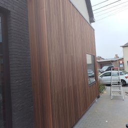 Kozik - Nowoczesna elewacja domu z drewnianą okładziną pionową, kontrastująca z ciemną, ceglaną ścianą. Widoczna aluminiowa drabina i piła tarczowa na kostce brukowej sugerują trwające prace wykończeniowe.