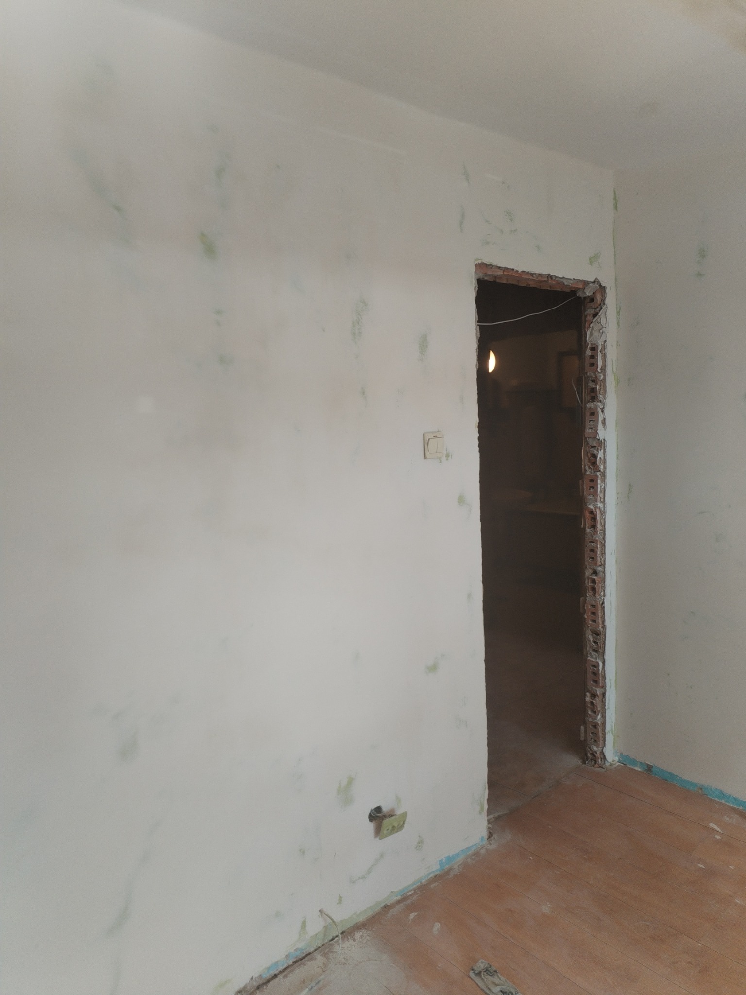 Ściana po wstępnym malowaniu z widocznymi smugami farby, przygotowane ościeże drzwiowe z odsłoniętą cegłą, drewniana podłoga zabezpieczona folią malarską.