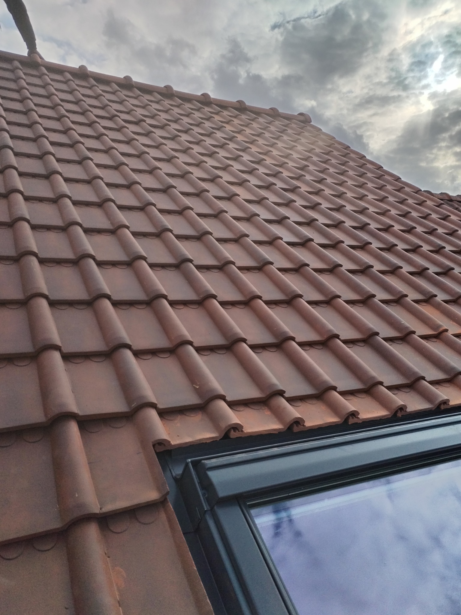 Nowy dach z brązowej dachówki ceramicznej z oknem dachowym, widok z dołu na tle pochmurnego nieba.