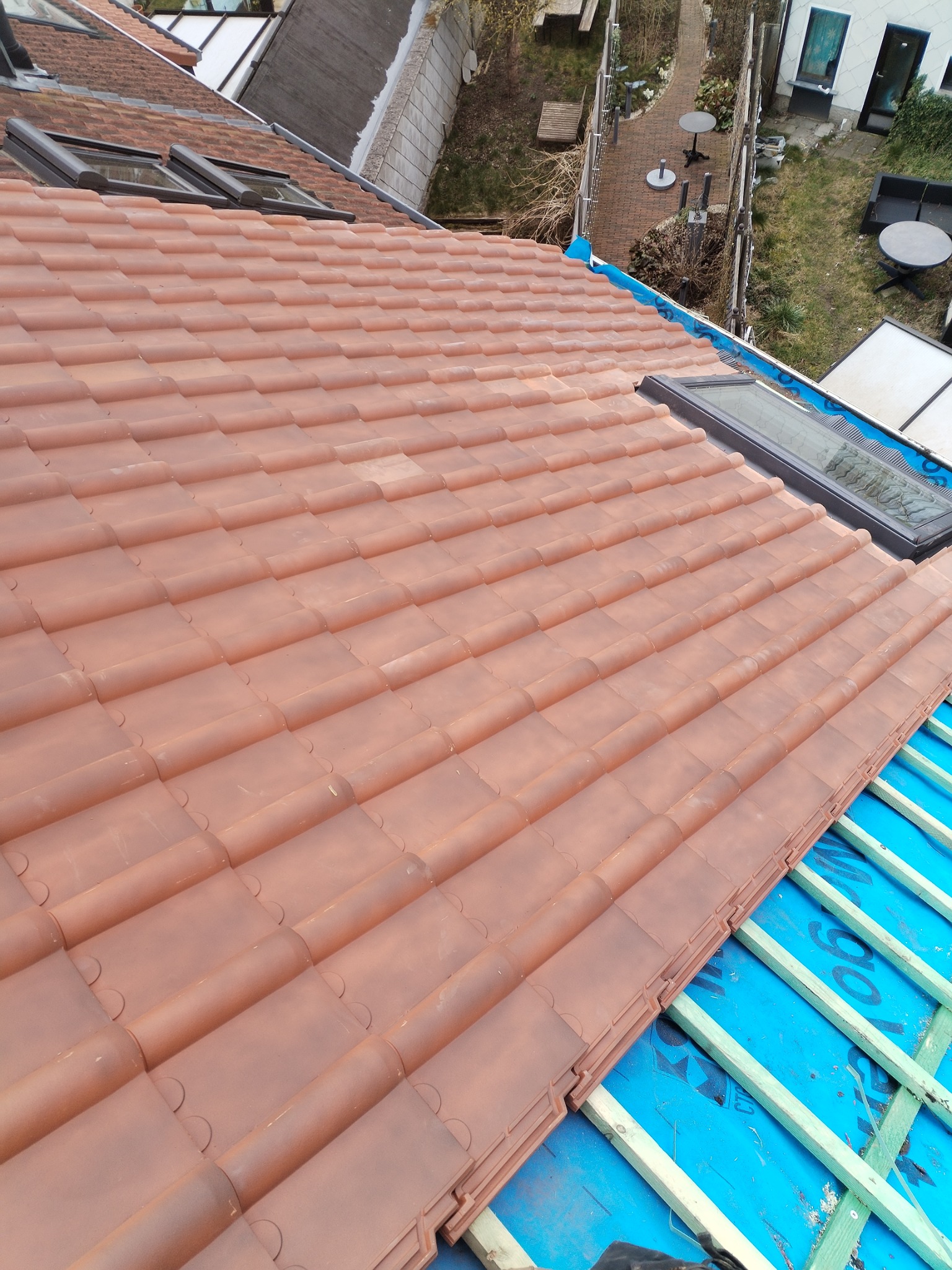 Nowy dach z ceramicznej dachówki w odcieniach terakoty, widoczne okna dachowe i fragment konstrukcji z niebieską membraną, ujęcie z góry na tle ogrodu.