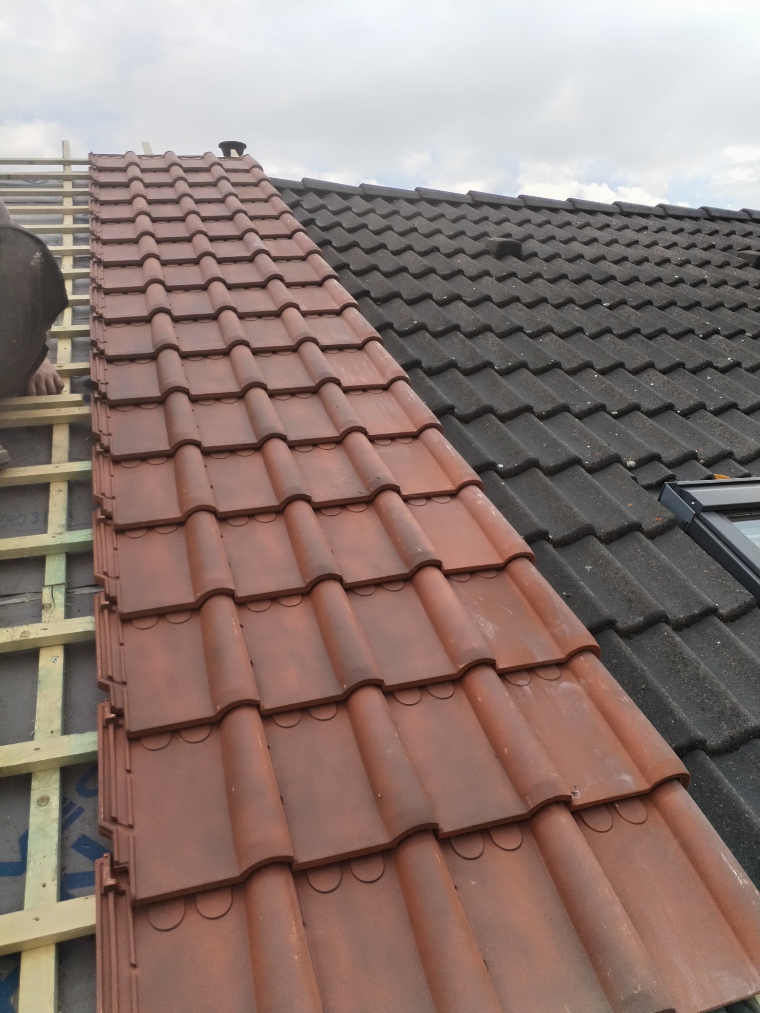 Układanie nowych, brązowych dachówek na dachu, widoczny fragment starego, czarnego pokrycia oraz drewniana konstrukcja łatowania.