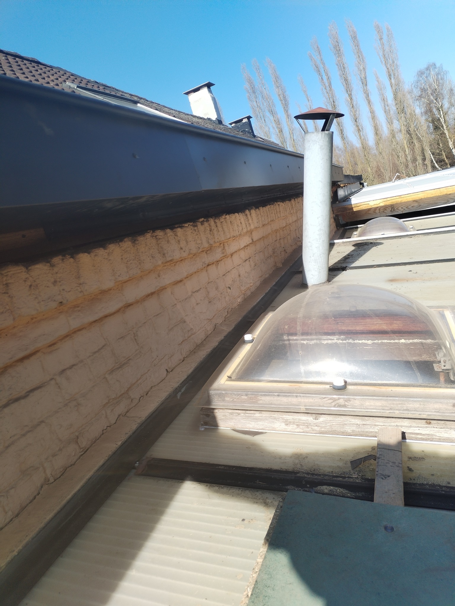 Widok z góry na część dachu z kominem, oknami dachowymi i ociepleniem ściany szczytowej imitującym cegłę, w tle wysokie drzewa bez liści.