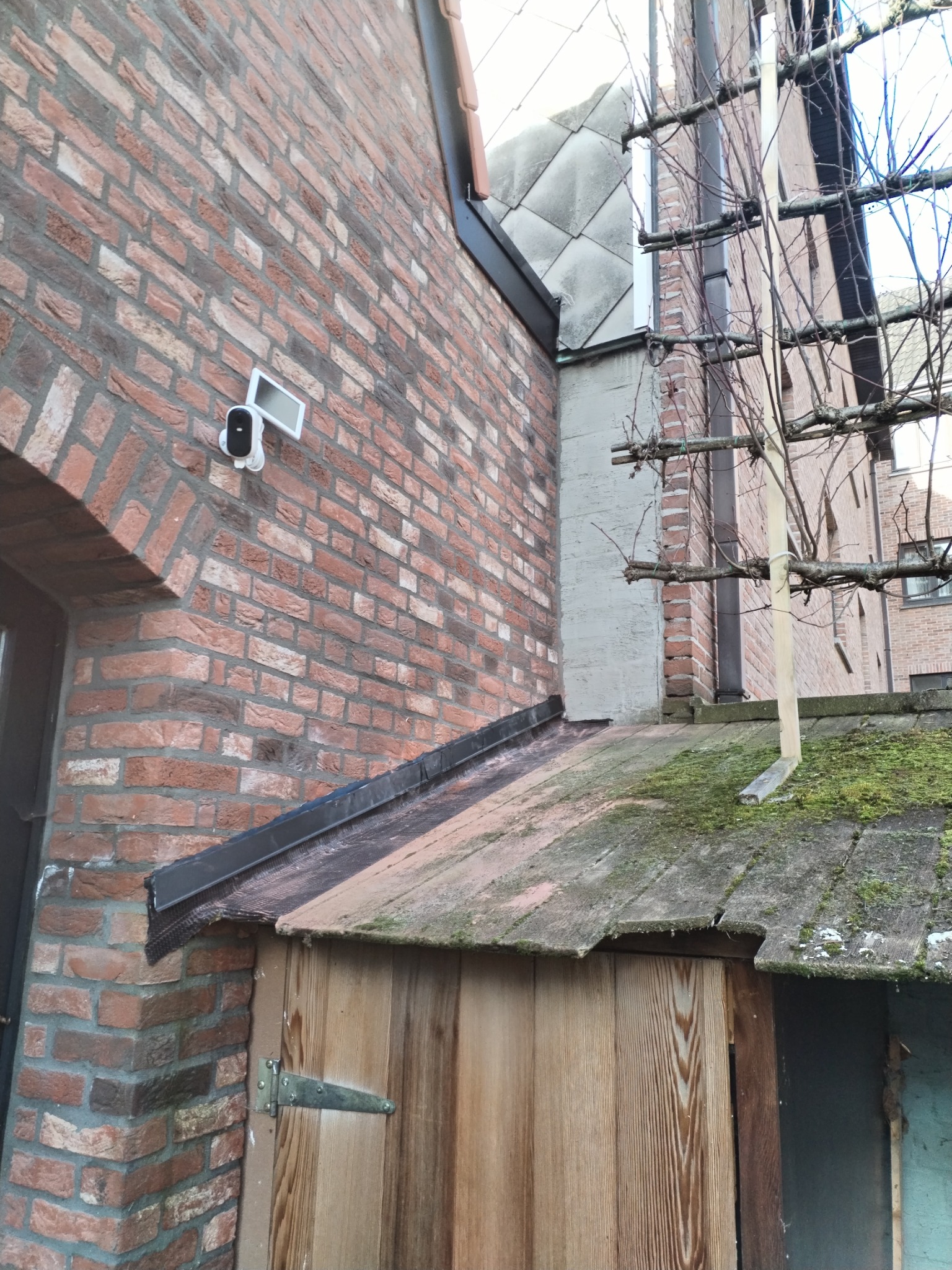 Czerwona ceglana ściana budynku z zamontowaną kamerą monitoringu, przylegająca do zadaszonej konstrukcji z mchem na dachu.