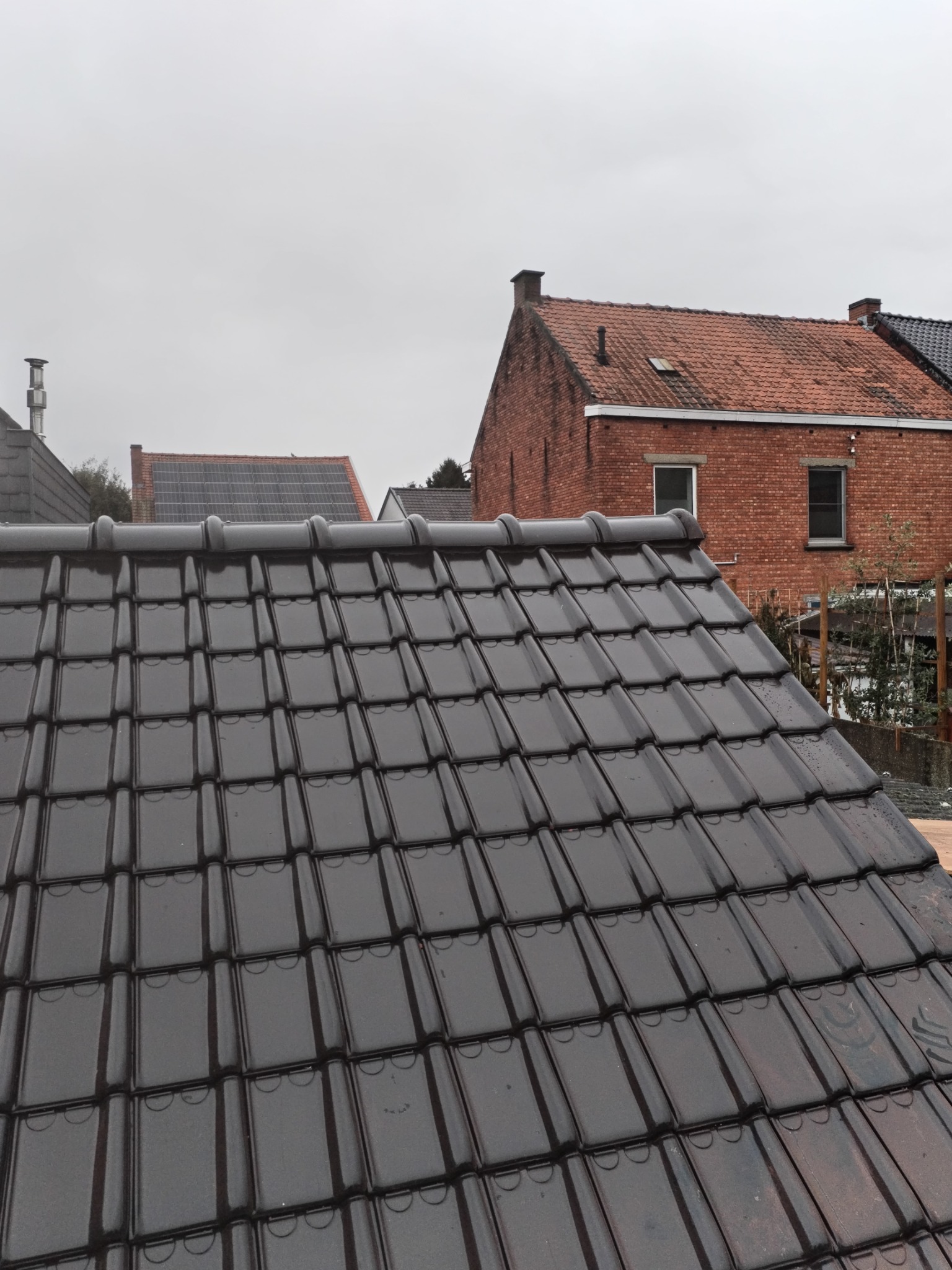 Błyszczące, ciemne dachówki ceramiczne na tle pochmurnego nieba i zabudowań mieszkalnych.