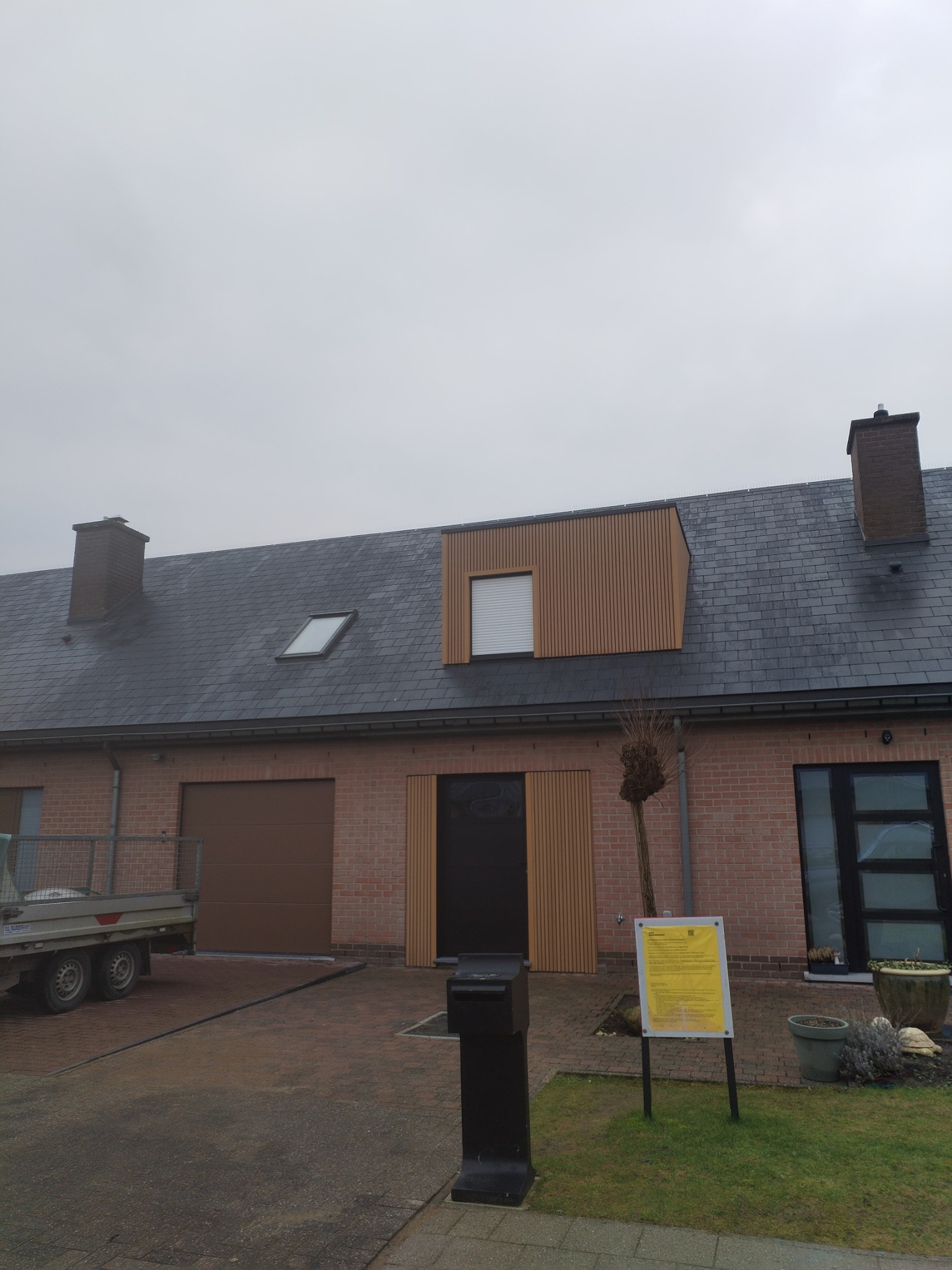 Dwukondygnacyjny dom z cegły z ciemnoszarym dachem z dachówki, oknem dachowym i drewnianą elewacją nad wejściem, widok z zewnątrz w pochmurny dzień.