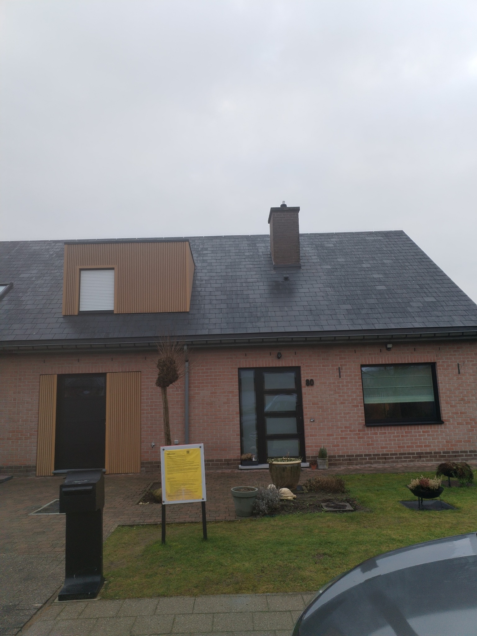 Dom jednorodzinny z ciemnoszarym dachem krytym łupkiem, ceglaną elewacją i nowoczesnymi elementami z drewnianych paneli pionowych. Na dachu widoczny komin. Na trawniku przed domem stoi tablica...