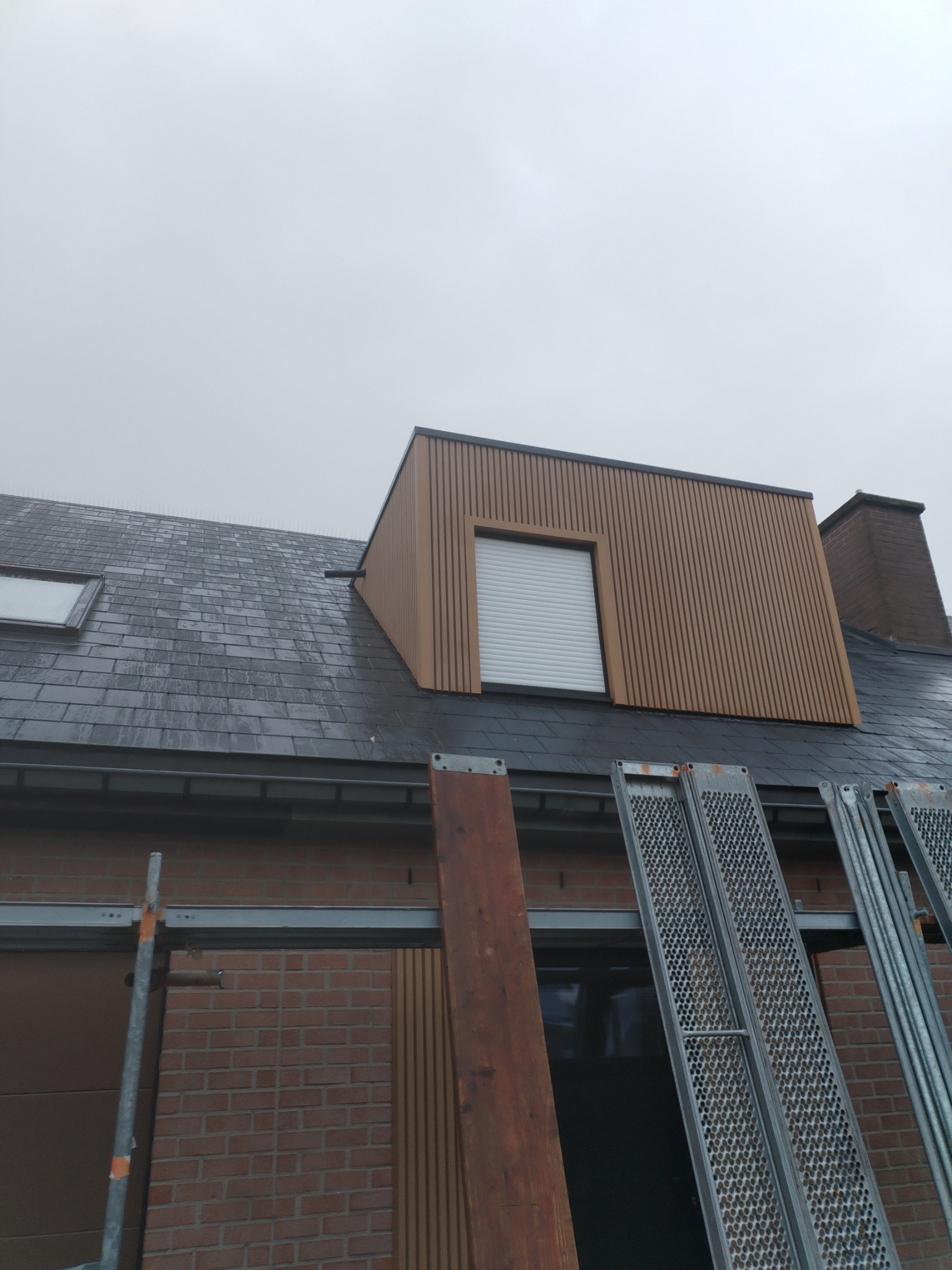 Nowo wykończona lukarna z brązową okładziną pionową, szara dachówka i rusztowanie na tle pochmurnego nieba.