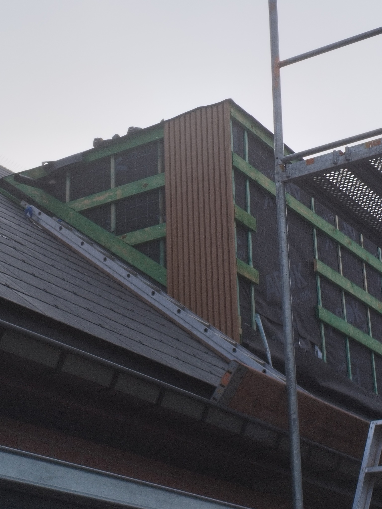 Konstrukcja dachu z łatami, membraną i częściowo zamontowaną brązową elewacją pionową, widoczna drabina i rusztowanie.