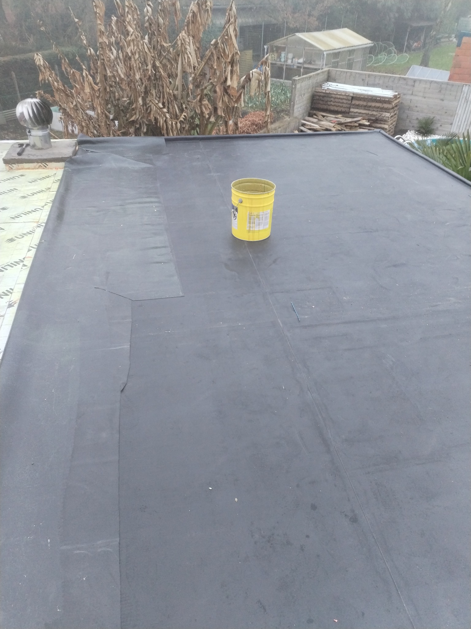 Płaski dach pokryty czarną papą z widocznymi łączeniami i łatami, na środku stoi żółte wiadro, w tle kominek wentylacyjny i zabudowania gospodarcze.