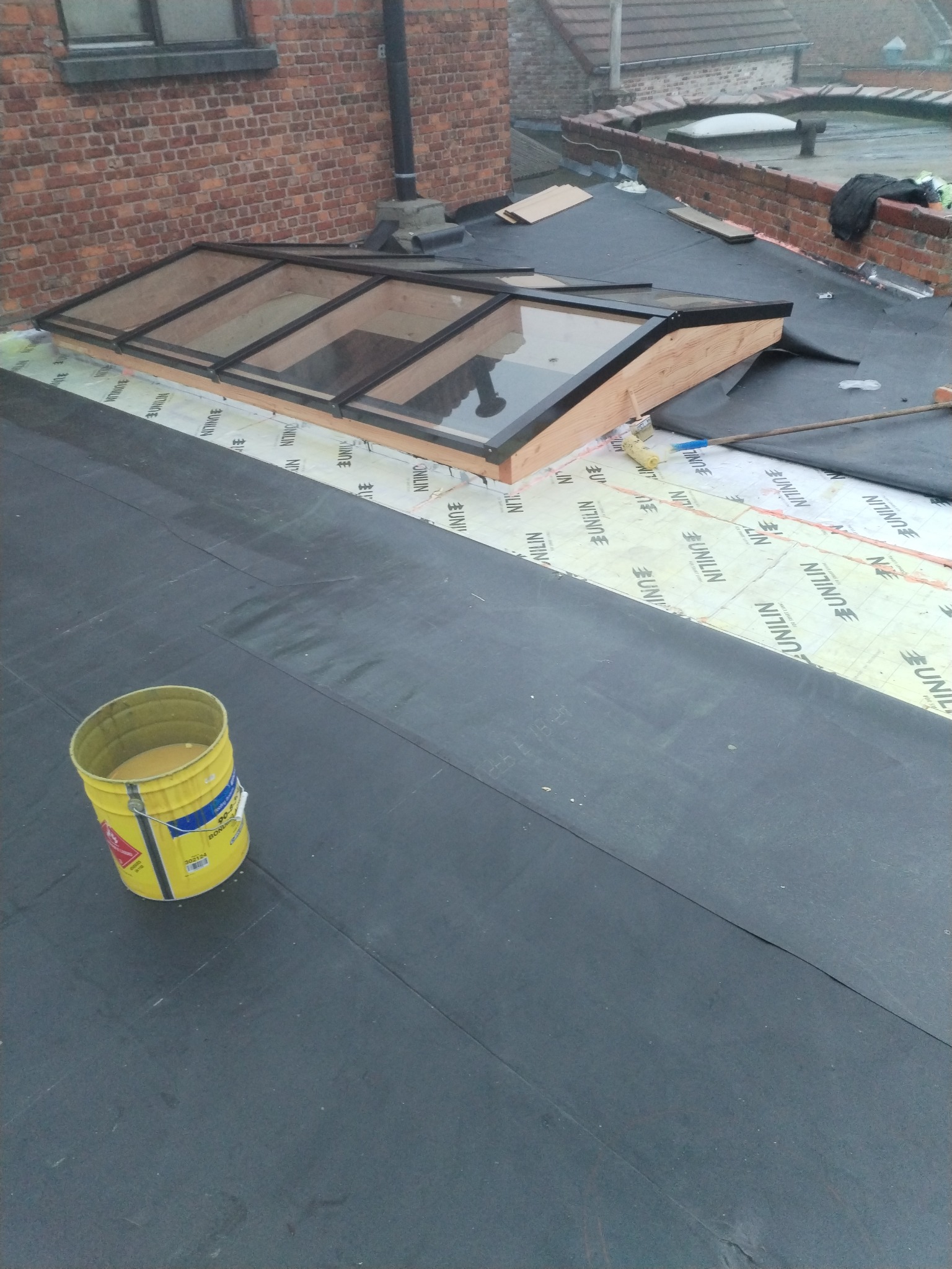 Montaż okna dachowego na dachu pokrytym papą, widoczny żółty pojemnik z klejem i rolka do rozprowadzania kleju na membranie izolacyjnej Unilin.