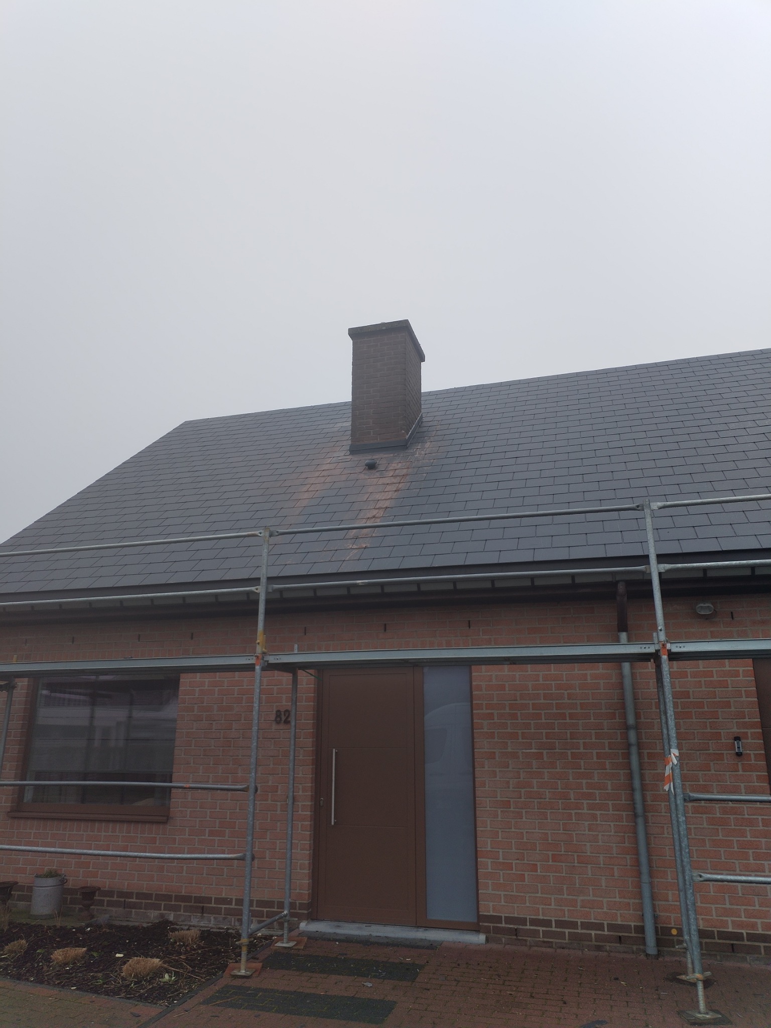 Dom jednorodzinny z nowym dachem z szarej dachówki, widoczne rusztowanie i komin z cegły, pochmurna pogoda.