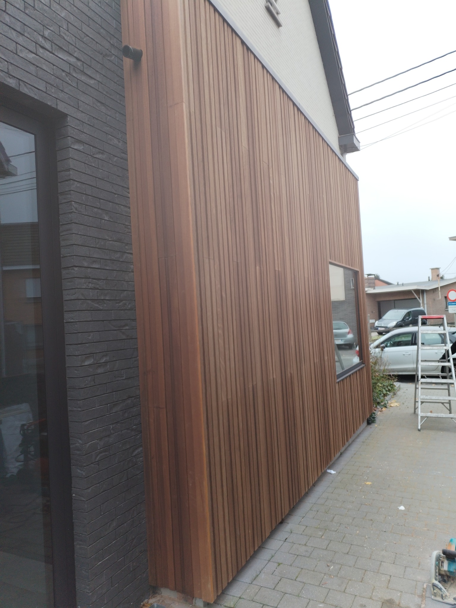 Nowoczesna elewacja budynku z drewnianą okładziną pionową w ciepłym odcieniu brązu, kontrastująca z ciemną, ceglaną ścianą obok okna. Na chodniku widoczna drabina i narzędzia.