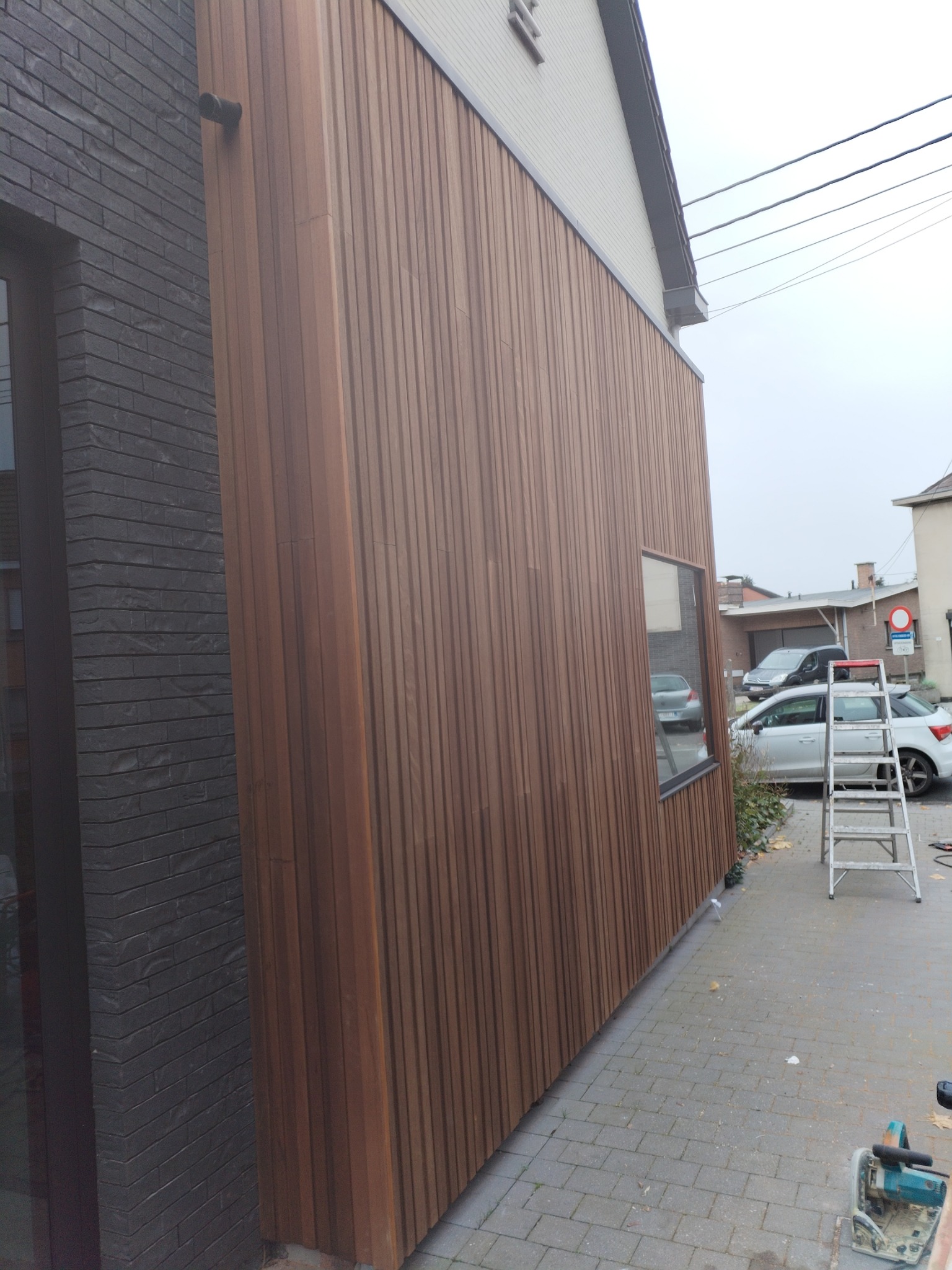 Nowoczesna elewacja domu z drewnianą okładziną pionową, kontrastująca z ciemną, ceglaną ścianą. Widoczna aluminiowa drabina i piła tarczowa na kostce brukowej sugerują trwające prace wykończeniowe.