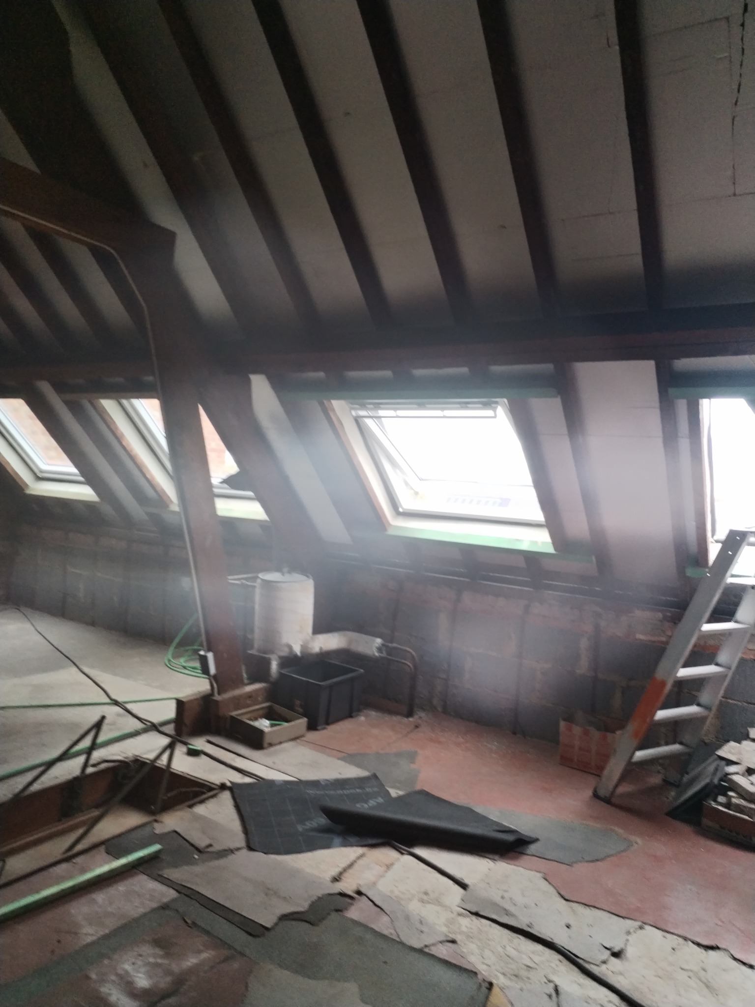 Poddasze w trakcie prac adaptacyjnych z widocznymi oknami dachowymi, drewnianą konstrukcją więźby dachowej, elementami instalacji, gruzem i drabiną opartą o ścianę.