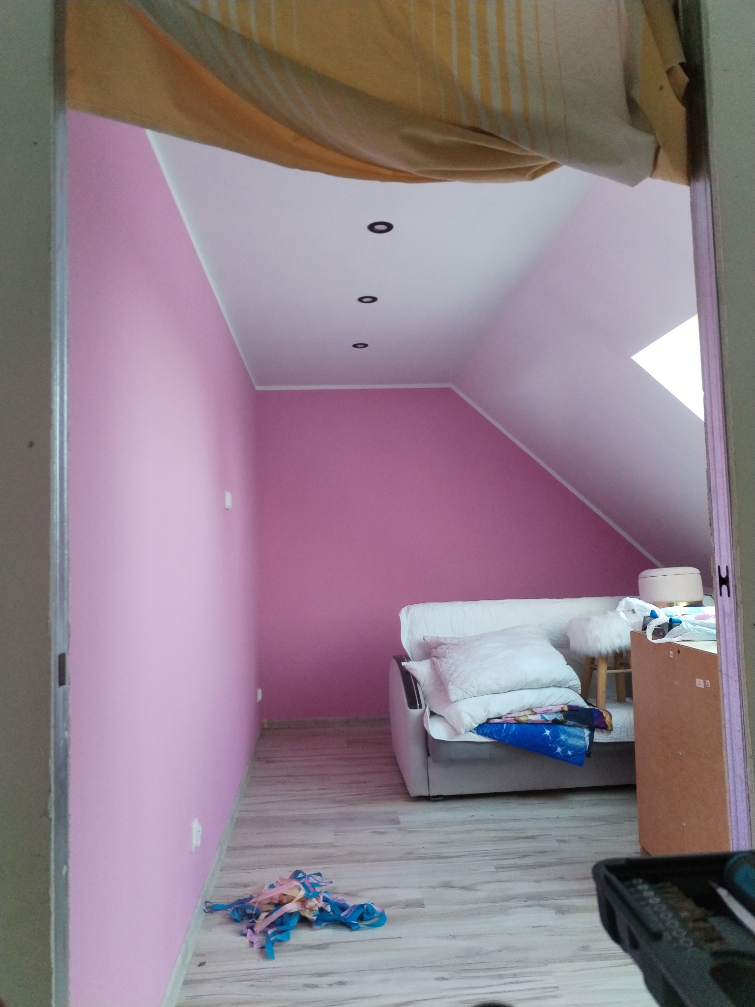 Pokój na poddaszu po malowaniu ścian na różowo, widoczne meble, okno dachowe i resztki taśmy malarskiej na podłodze. Oświetlenie punktowe sufitowe.