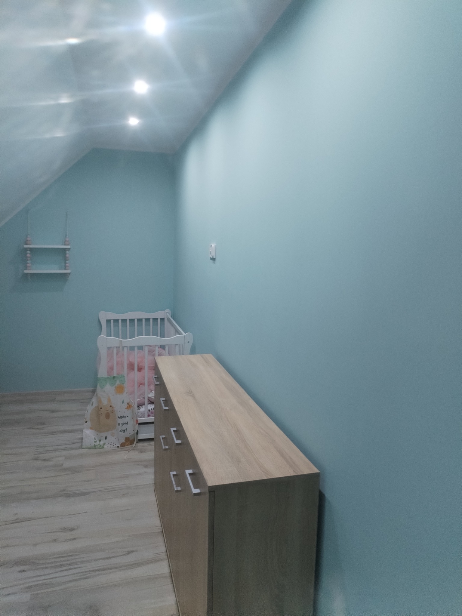 Niebieski pokój dziecięcy na poddaszu z drewnianą komodą, łóżeczkiem i półką wiszącą. Ściany pomalowane na jednolity kolor, podłoga laminowana imitująca drewno.