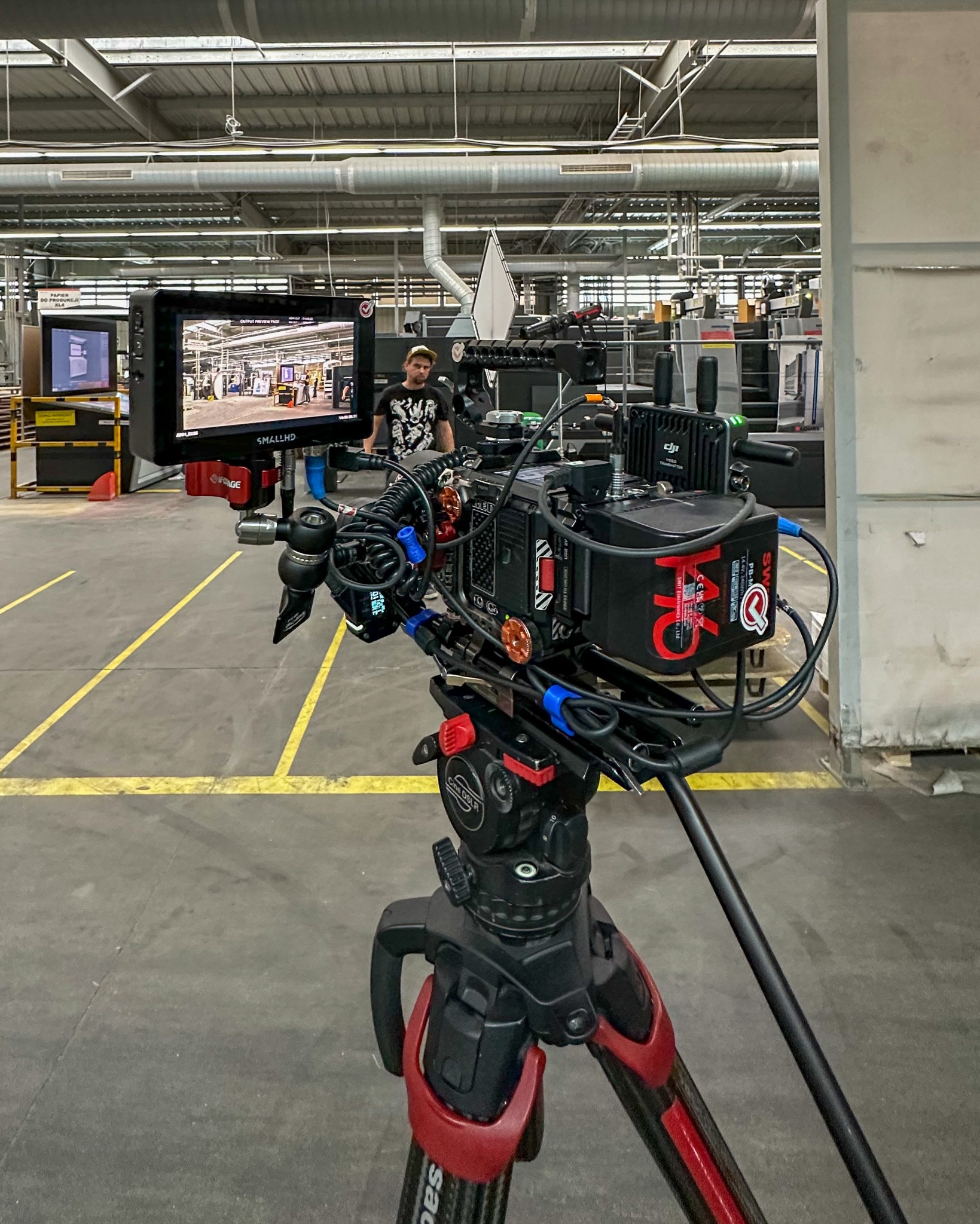 Profesjonalna kamera filmowa na statywie, monitor podglądowy pokazuje wnętrze hali produkcyjnej, w tle operator w czarnej koszulce.