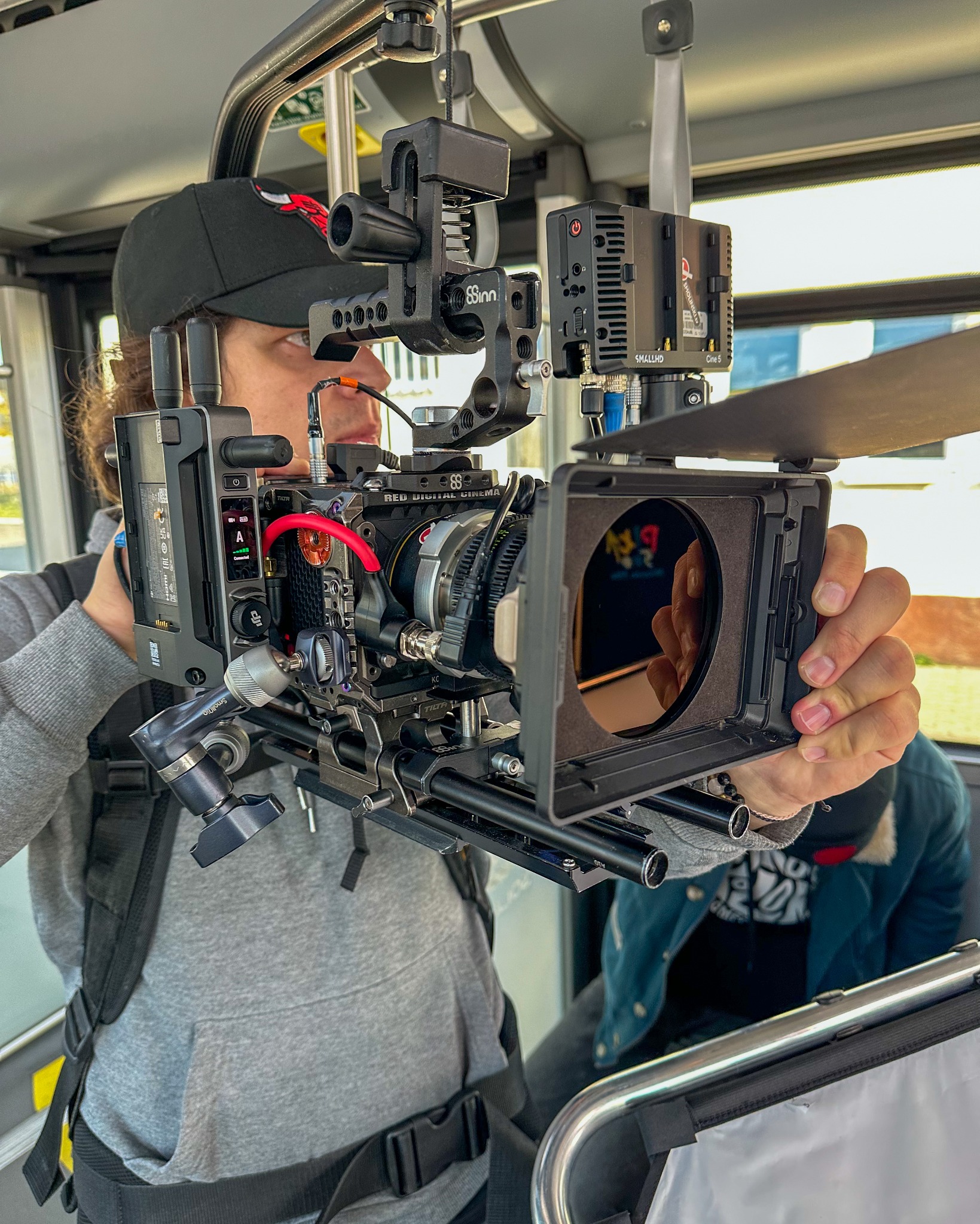 Operator z profesjonalną kamerą RED Digital Cinema z zamontowanymi akcesoriami SmallHD i Tilta, w trakcie filmowania, widoczny kadr z odbiciem w obiektywie.