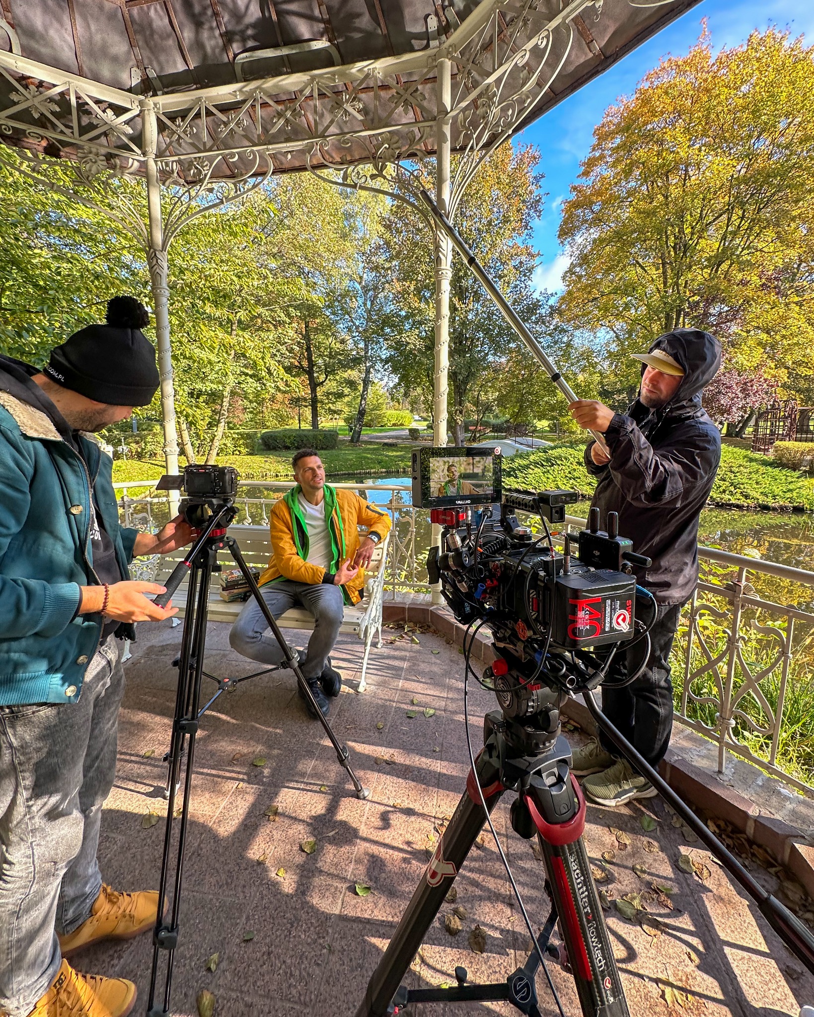 Ekipa filmowa kręci ujęcie z aktorem siedzącym w altanie w parku, widoczny profesjonalny sprzęt filmowy i mikrofon na tyczce.