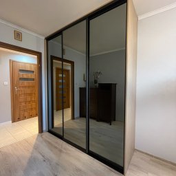 MARBEL.PL - Szafa wnękowa z przesuwnymi, lustrzanymi drzwiami, w jasnym odcieniu drewna, z czarnymi obramowaniami, odbijająca wnętrze pokoju z komodą i kwiatami, na tle jasnych ścian i drewnianej podłogi.