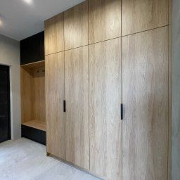 MARBEL.PL - Wysoka szafa wnękowa z jasnego drewna z czarnymi, podłużnymi uchwytami, połączona z otwartą garderobą z wieszakami i szafką w kolorze czarnym.