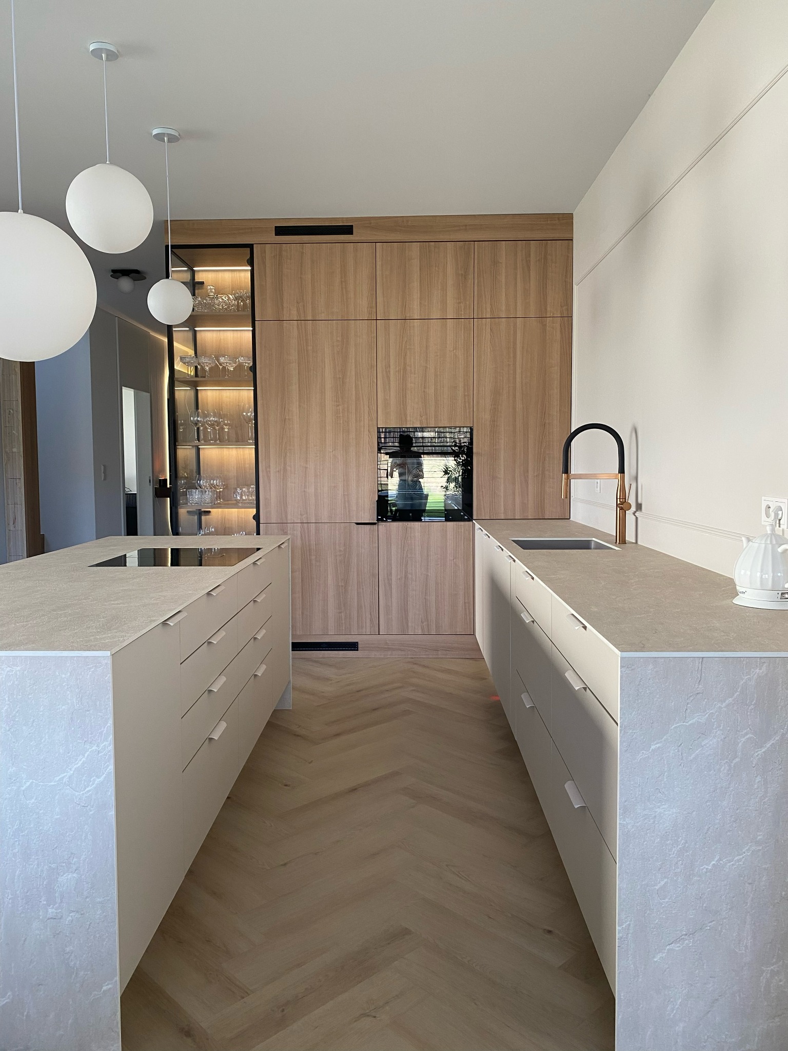 Nowoczesna kuchnia z dwoma wyspami, drewnianymi szafkami i podłogą w jodełkę. Widoczna przeszklona szafka z oświetleniem i szklanymi naczyniami oraz czarno-złota bateria kuchenna.