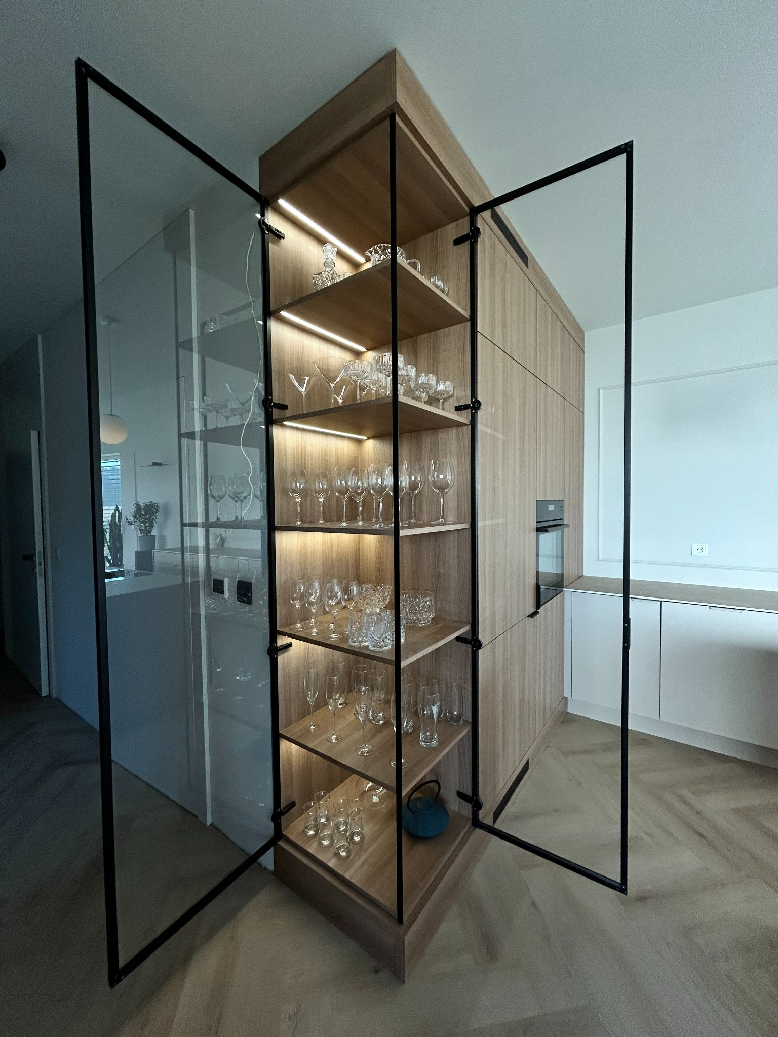Drewniana szafa z przeszklonymi drzwiami w czarnych ramach, podświetlona, wypełniona szkłem dekoracyjnym i kieliszkami, w tle biała kuchnia.