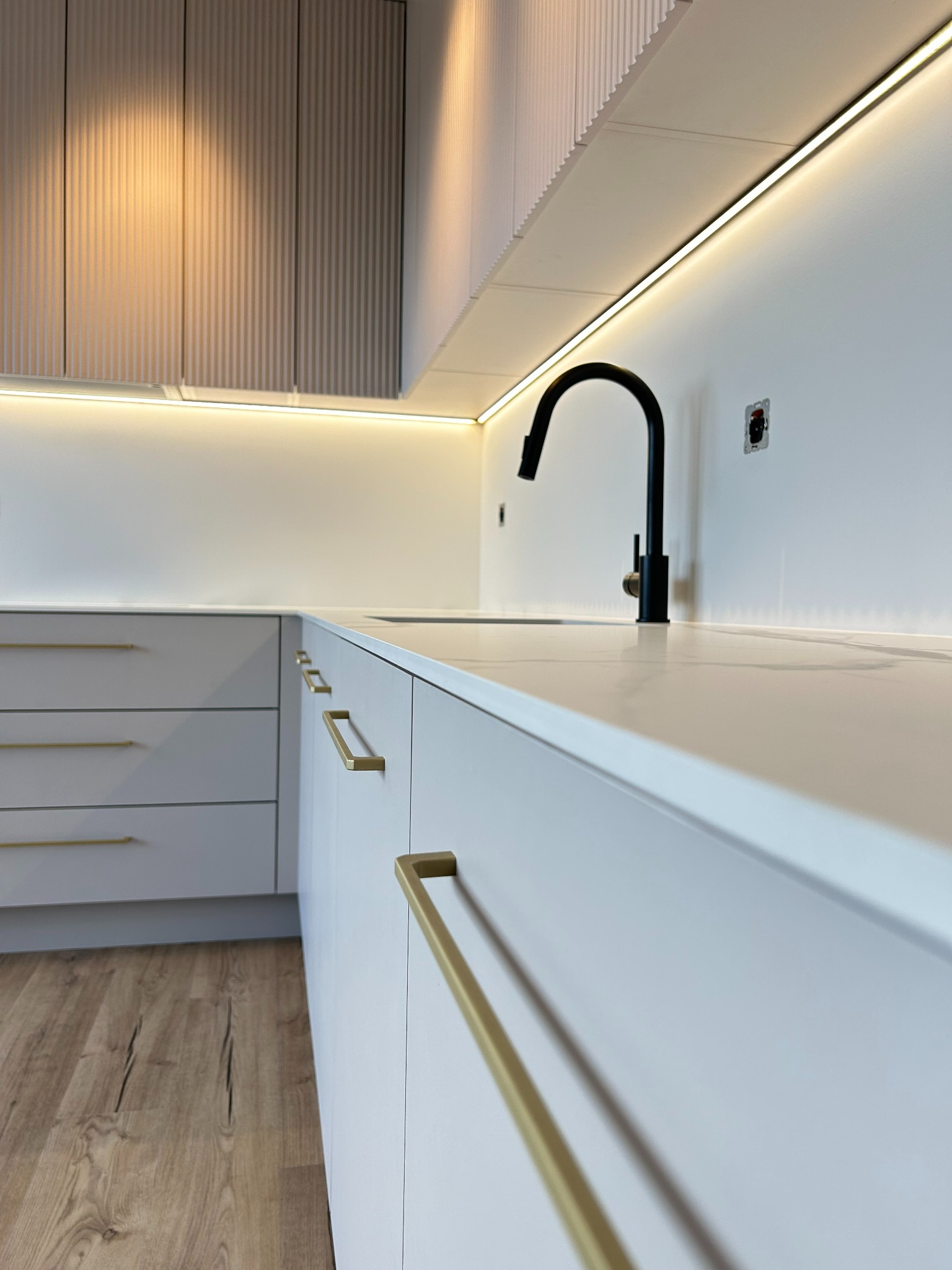 Nowoczesna kuchnia z białymi frontami, złotymi uchwytami i czarną baterią, oświetlenie LED podszafkowe, drewniana podłoga.
