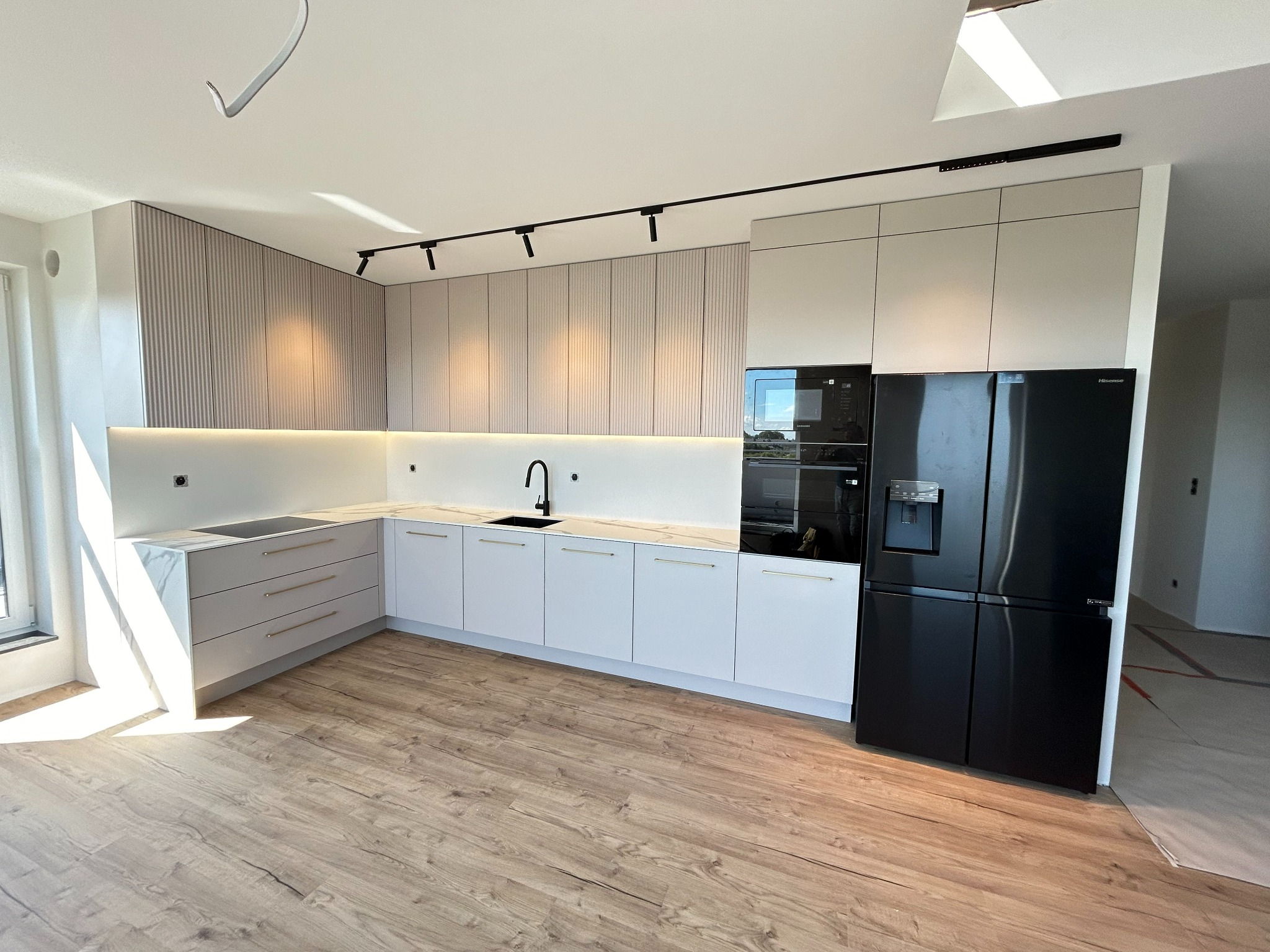 Nowoczesna kuchnia z frontami w odcieniach bieli i beżu, z czarnym oświetleniem punktowym i dużą czarną lodówką side-by-side, jasna podłoga imitująca drewno, widoczne elementy wykończeniowe.