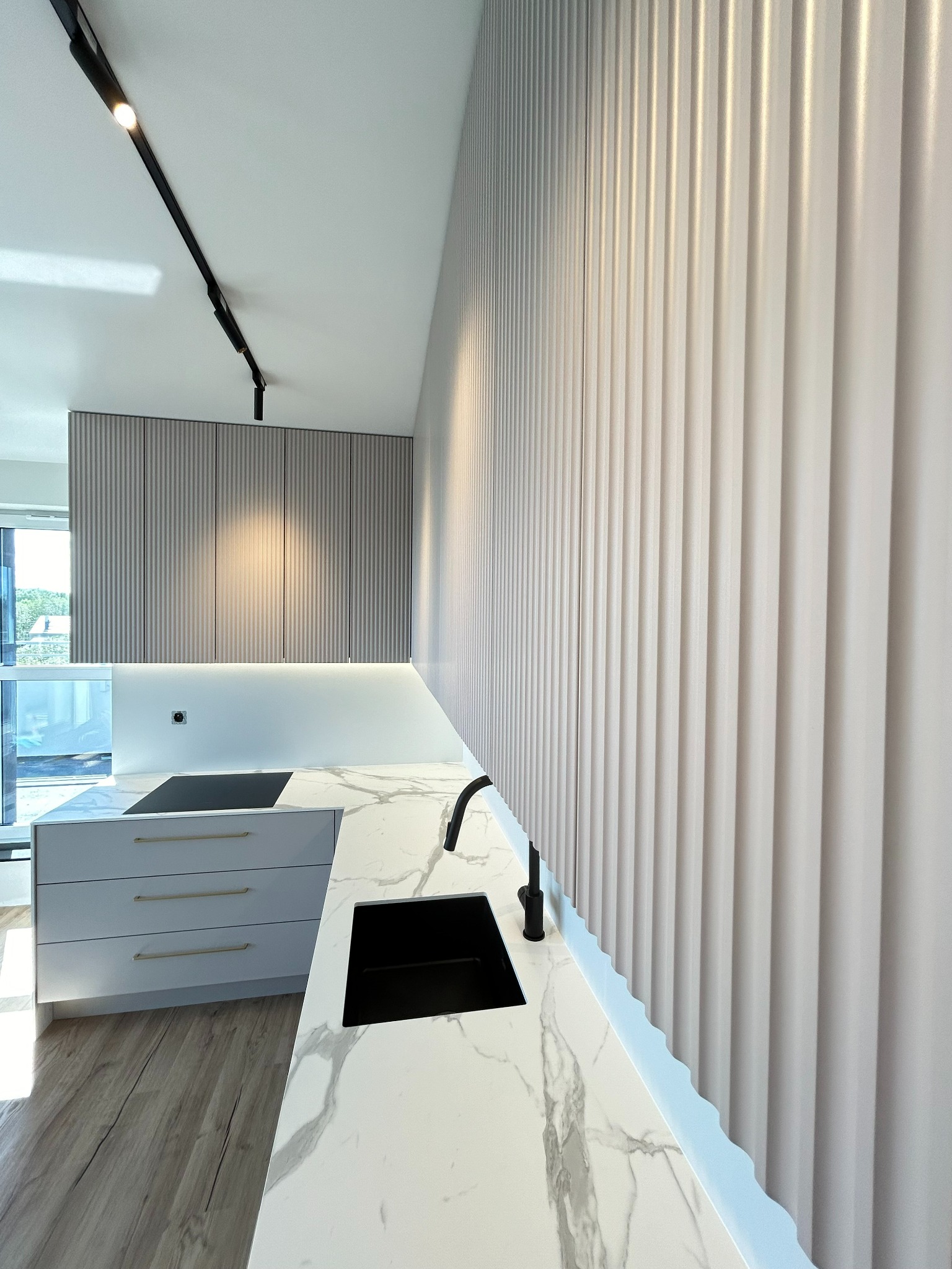 Nowoczesna kuchnia z frontami szafek w pionowe prążki, blat z białego marmuru z czarnym zlewem i baterią, oświetlenie podszafkowe LED, drewniana podłoga.