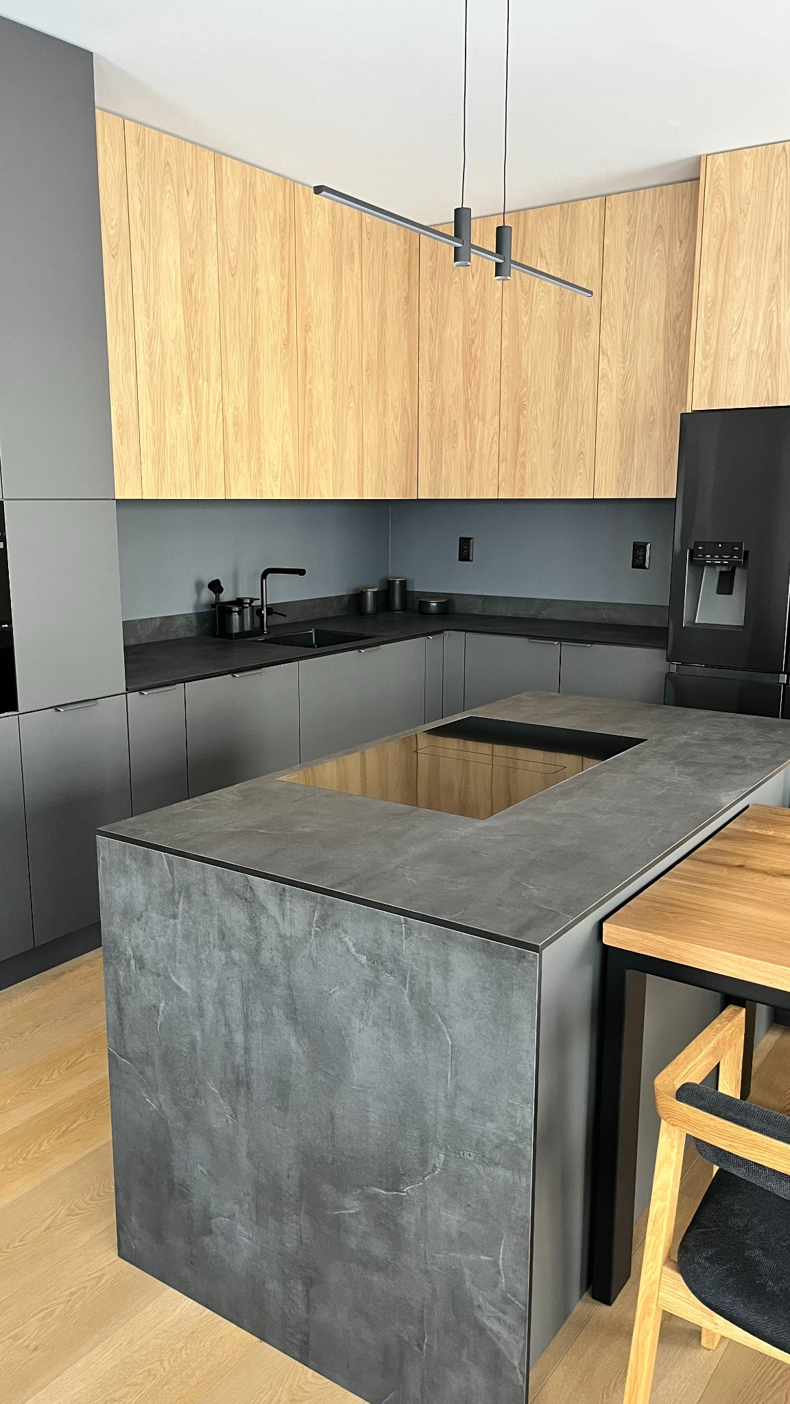 Nowoczesna kuchnia z szarymi dolnymi szafkami i drewnianymi górnymi, wyspa z blatem imitującym beton i drewnianym wnęką, minimalistyczna lampa sufitowa.