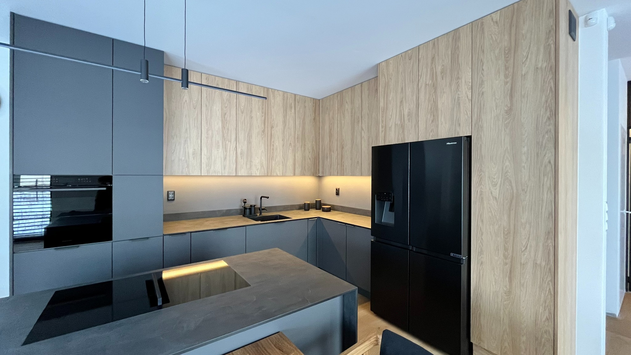 Nowoczesna kuchnia z szarymi frontami szafek dolnych i drewnopodobnymi górnymi, czarny sprzęt AGD, oświetlenie LED pod szafkami, szary blat i wyspa z czarną płytą indukcyjną.