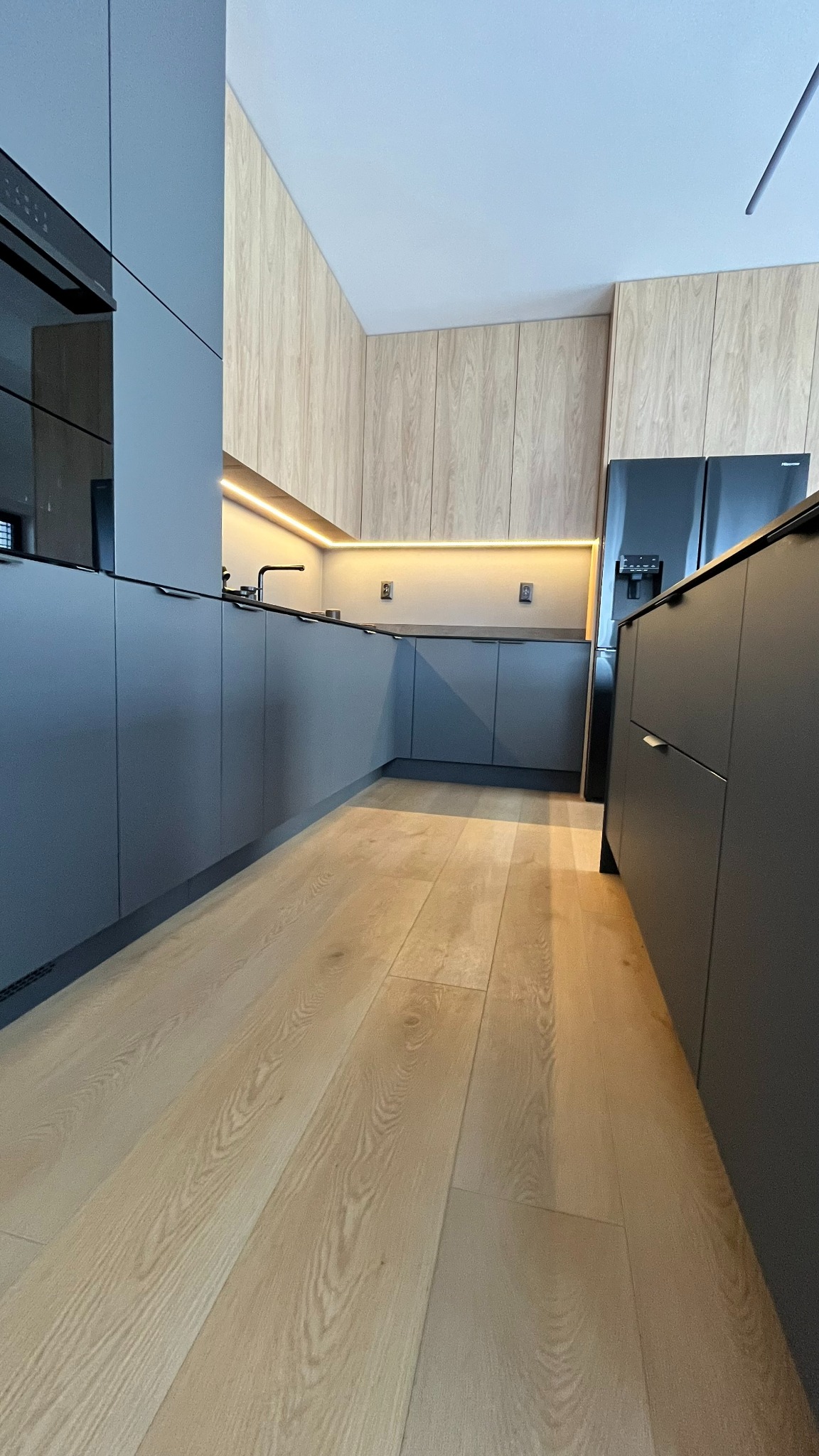 Nowoczesna kuchnia z szarymi frontami i drewnianymi panelami ściennymi, oświetlenie LED pod szafkami wiszącymi, jasna drewniana podłoga.