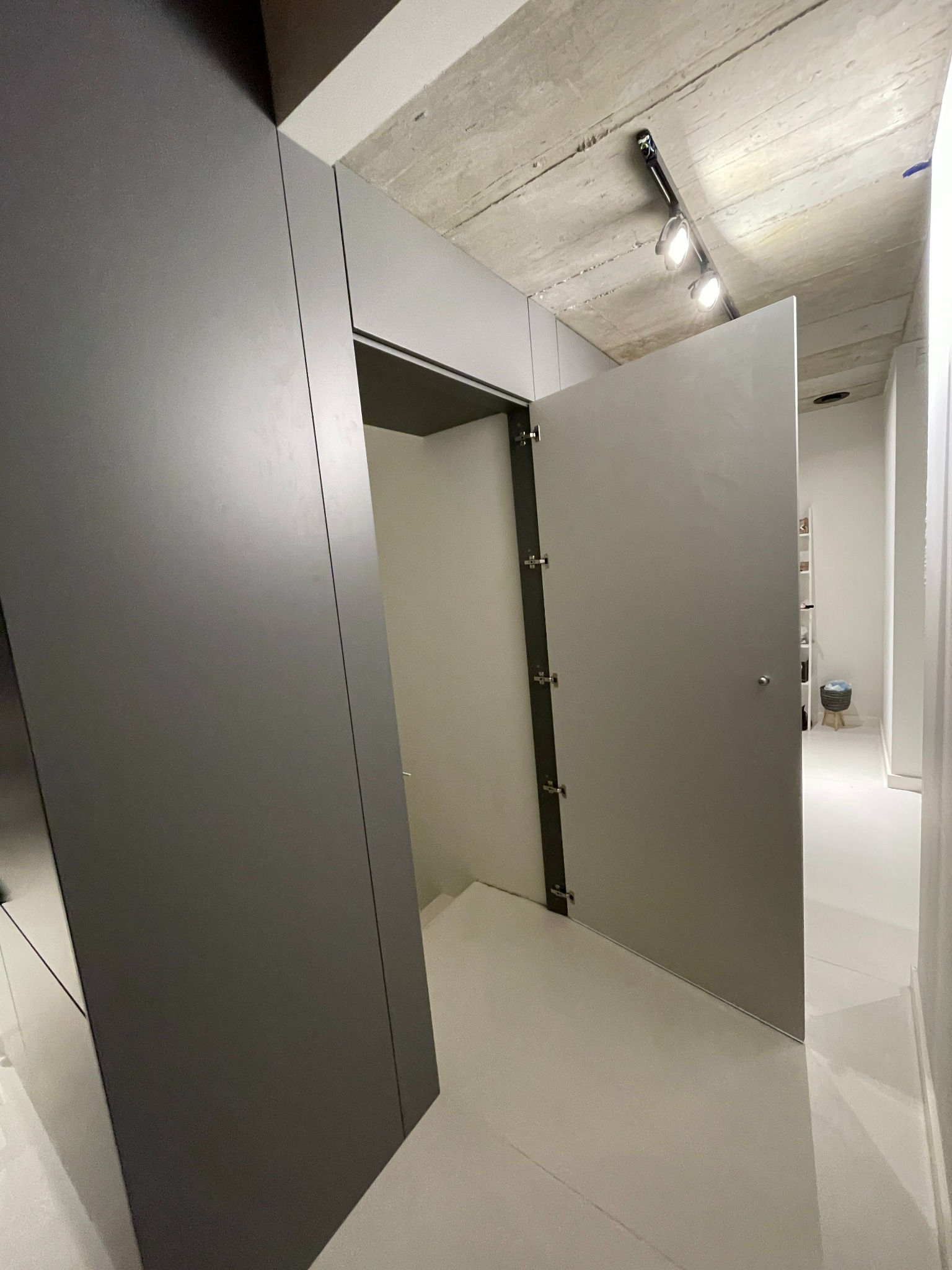 Minimalistyczna szafa wnękowa z szarymi frontami, otwierana do wewnątrz, widoczna na tle betonowego sufitu i jasnej podłogi w nowoczesnym wnętrzu.