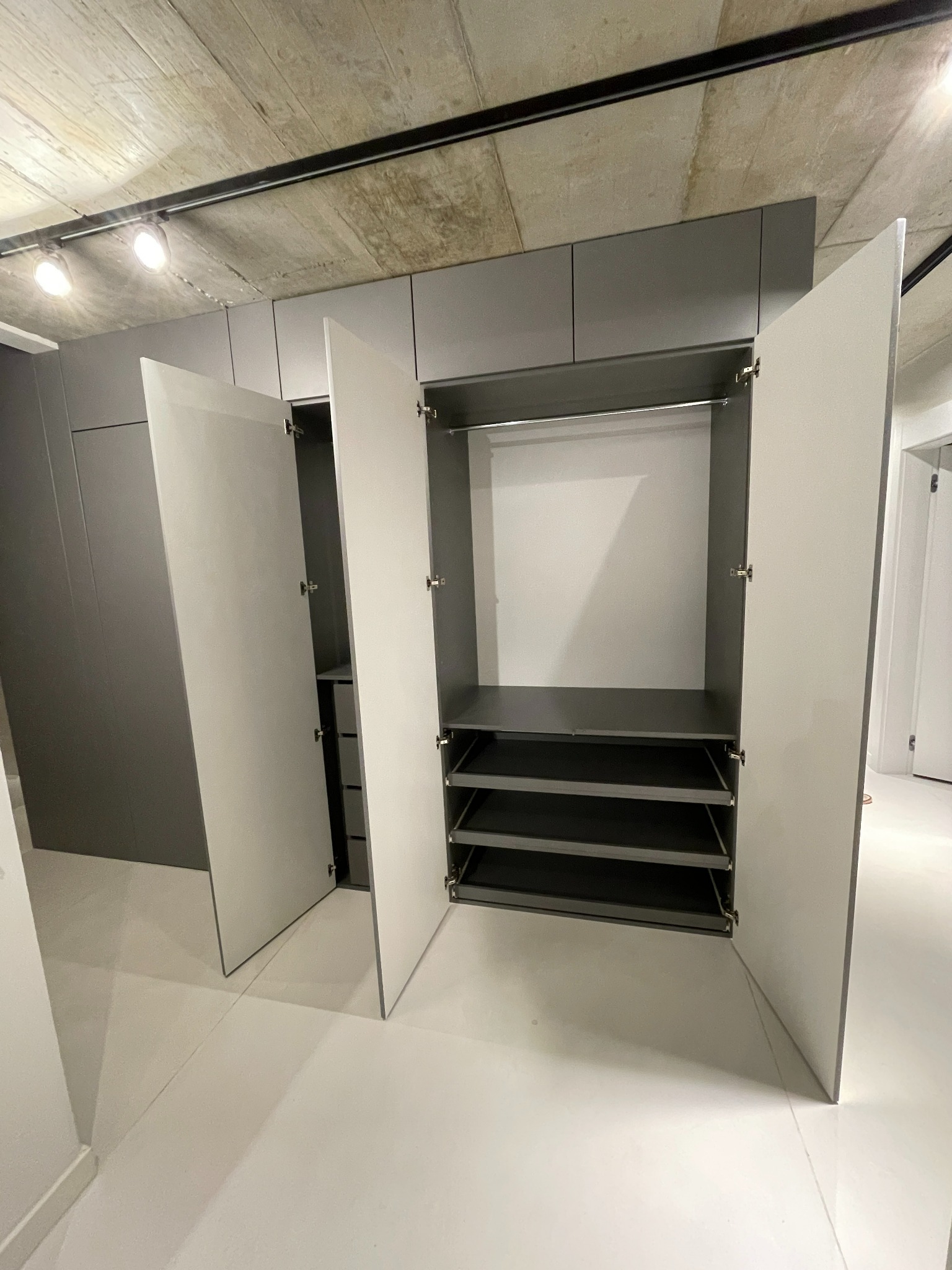 Szara szafa wnękowa z białymi drzwiami, otwarte drzwi ukazują półki i drążek, surowy betonowy sufit z oświetleniem punktowym.