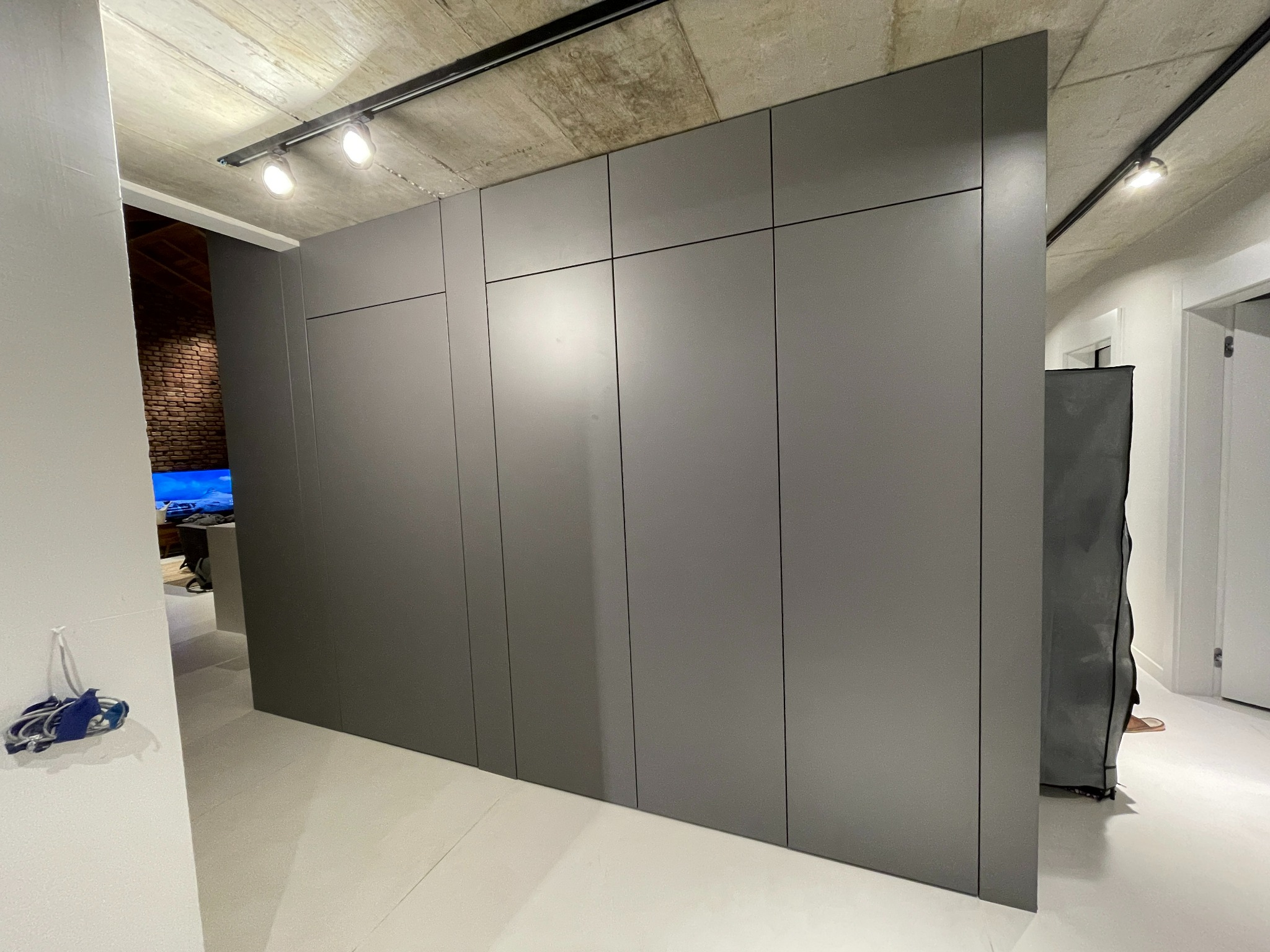 Szara szafa wnękowa z gładkimi frontami, oświetlenie szynowe na surowym betonowym suficie, widok na ceglaną ścianę w sąsiednim pomieszczeniu.