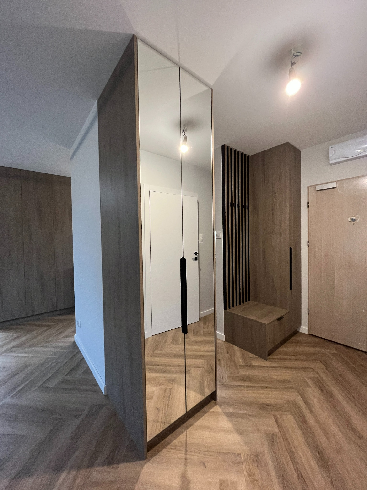 Nowoczesna szafa wnękowa z lustrzanymi drzwiami przesuwnymi, widoczna w przedpokoju z drewnianą podłogą w jodełkę i elementami zabudowy z lameli.