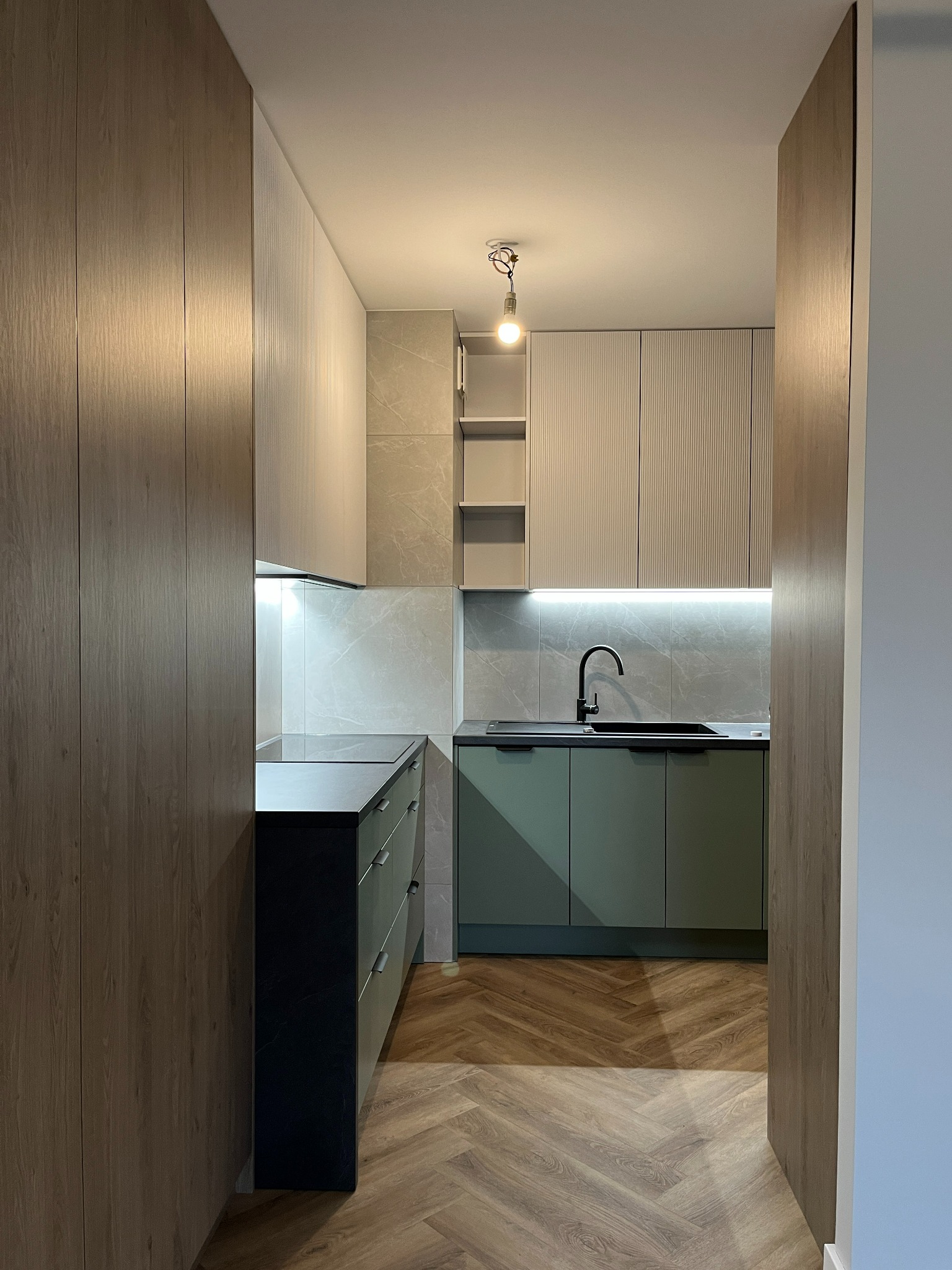 Nowoczesna kuchnia z zielonymi szafkami, czarnym zlewem i blatem, marmurowymi płytkami na ścianie i parkietem w jodełkę, oświetlona wiszącą żarówką i listwą LED pod szafkami.