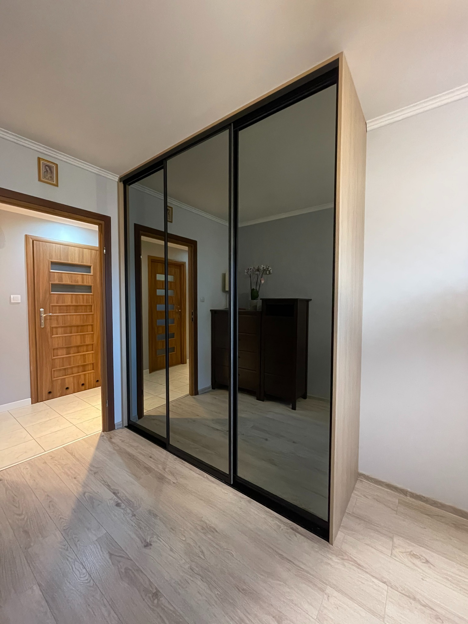 Szafa wnękowa z przesuwnymi, lustrzanymi drzwiami, w jasnym odcieniu drewna, z czarnymi obramowaniami, odbijająca wnętrze pokoju z komodą i kwiatami, na tle jasnych ścian i drewnianej podłogi.