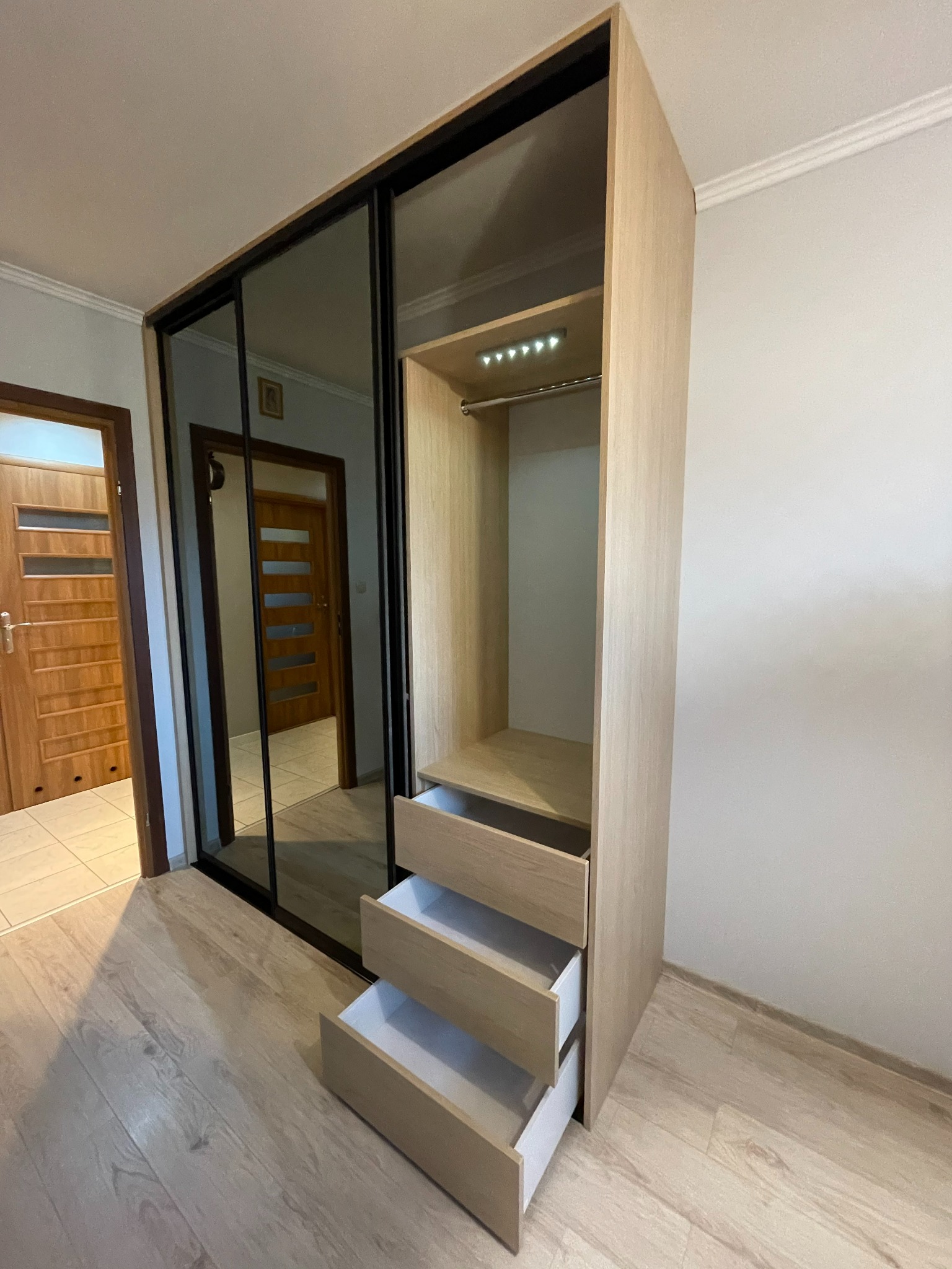 Jasna szafa wnękowa z przesuwnymi drzwiami z lustrami, otwartymi szufladami i oświetleniem LED wewnątrz.