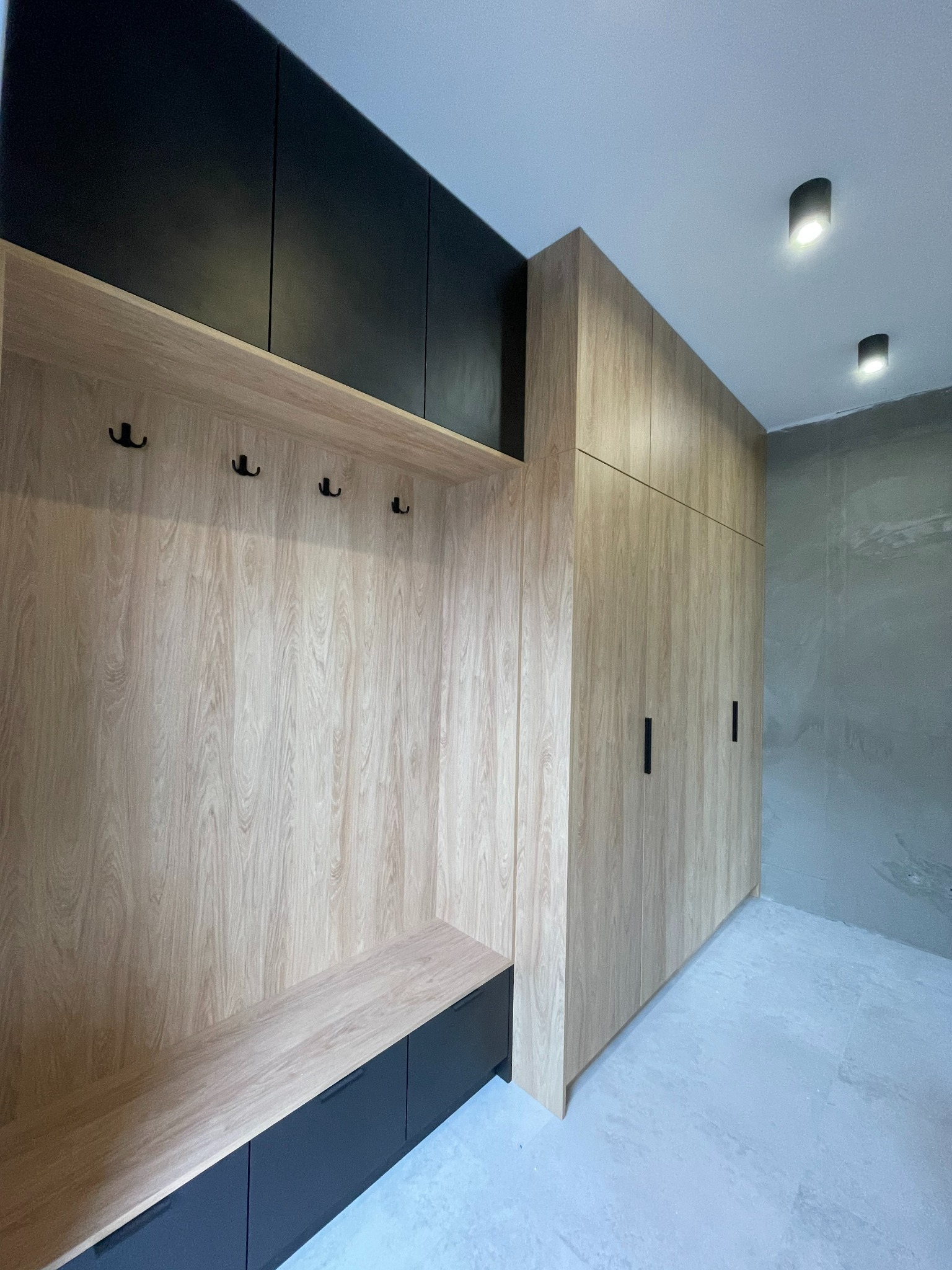 Wnęka wejściowa z drewnianymi panelami, czarnymi wieszakami i szafkami pod siedziskiem, obok wysoka szafa z drewnianymi drzwiami i czarnymi uchwytami.