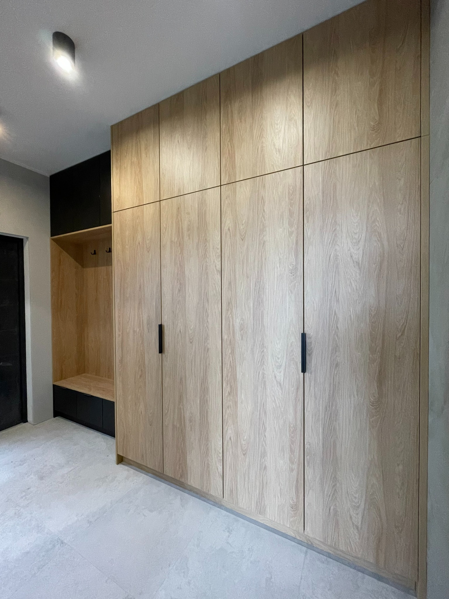 Wysoka szafa wnękowa z jasnego drewna z czarnymi, podłużnymi uchwytami, połączona z otwartą garderobą z wieszakami i szafką w kolorze czarnym.