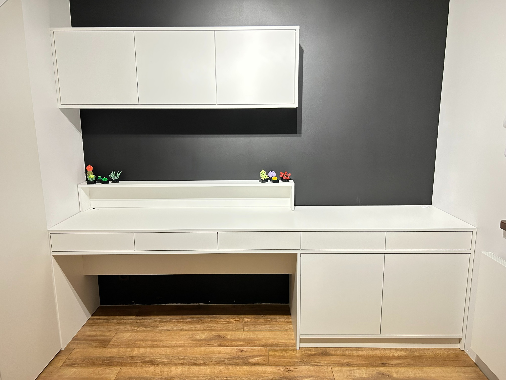 Białe biurko na wymiar z szafkami wiszącymi na tle szarej ściany, ozdobione miniaturowymi roślinami w doniczkach.