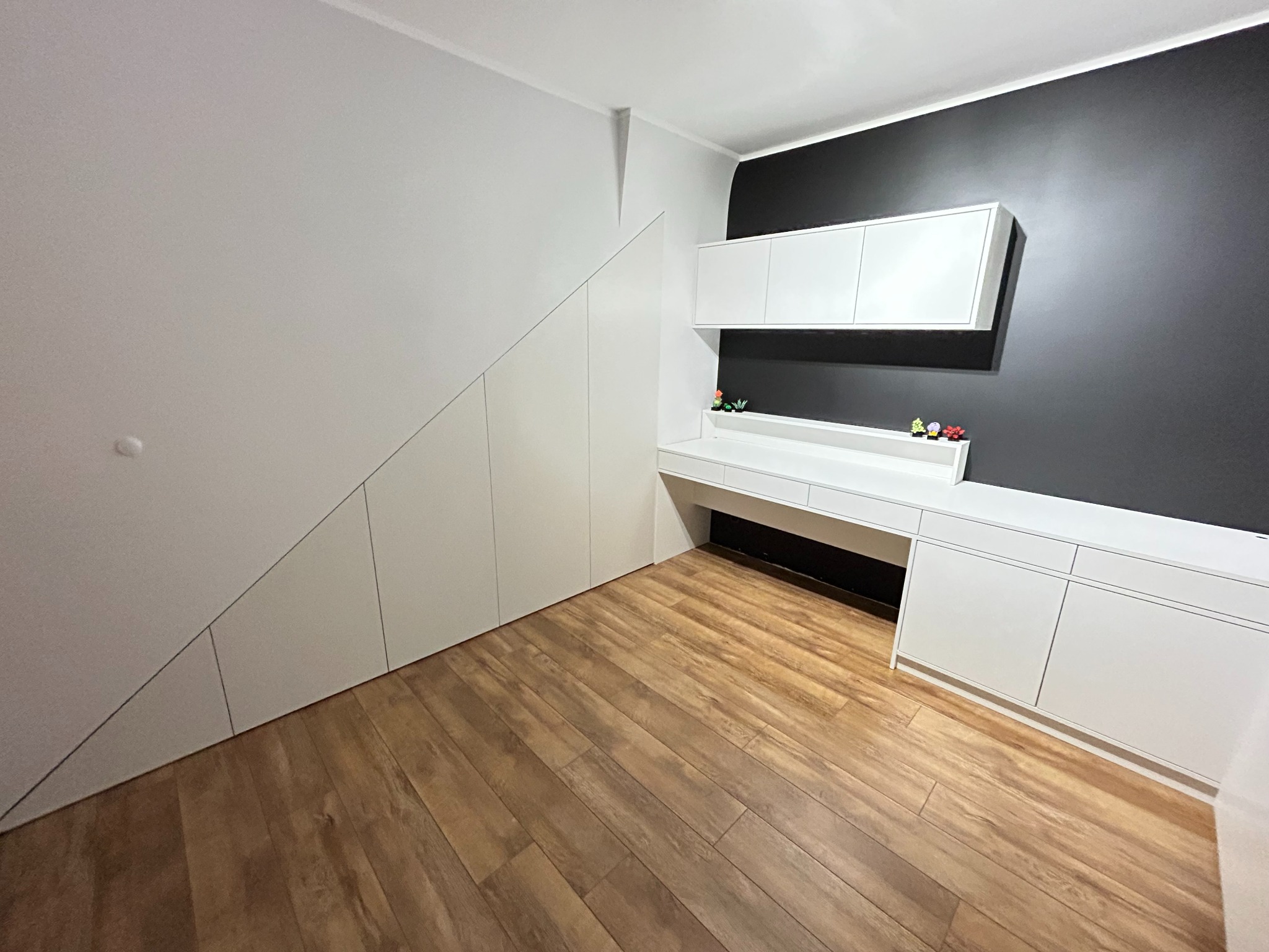 Biała zabudowa pod skosem z szafkami i biurkiem, po prawej szafka pod telewizor na tle czarnej ściany, drewniana podłoga.