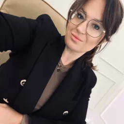 Portret kobiety w okularach i czarnej marynarce, siedzącej na krześle w biurze w Głogowie. Widoczny zegarek na ręce i złoty łańcuszek z zawieszkami.