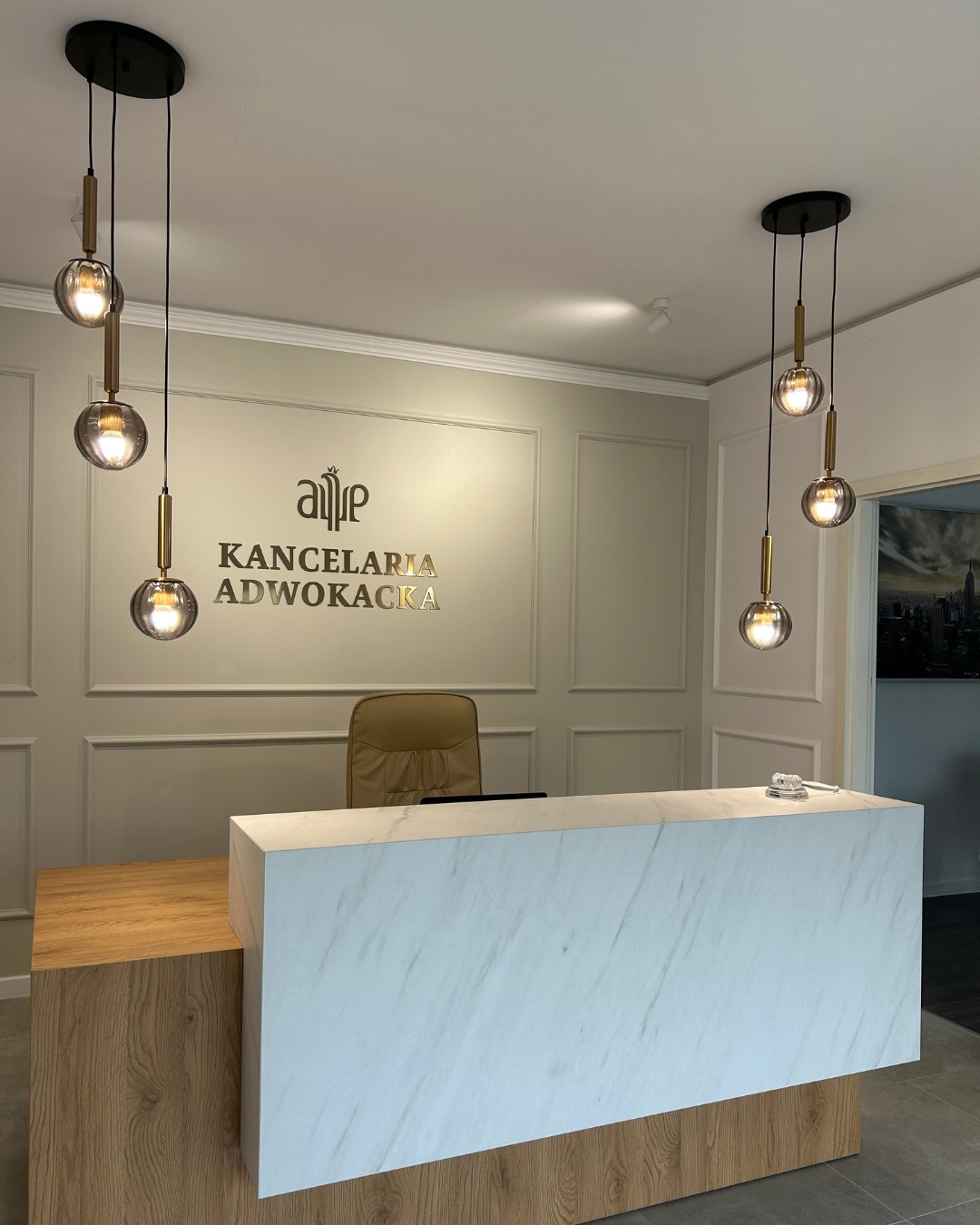 Recepcja w nowoczesnej kancelarii z marmurowym blatem, drewnianym wykończeniem i designerskimi lampami wiszącymi, w tle logo kancelarii na ścianie.