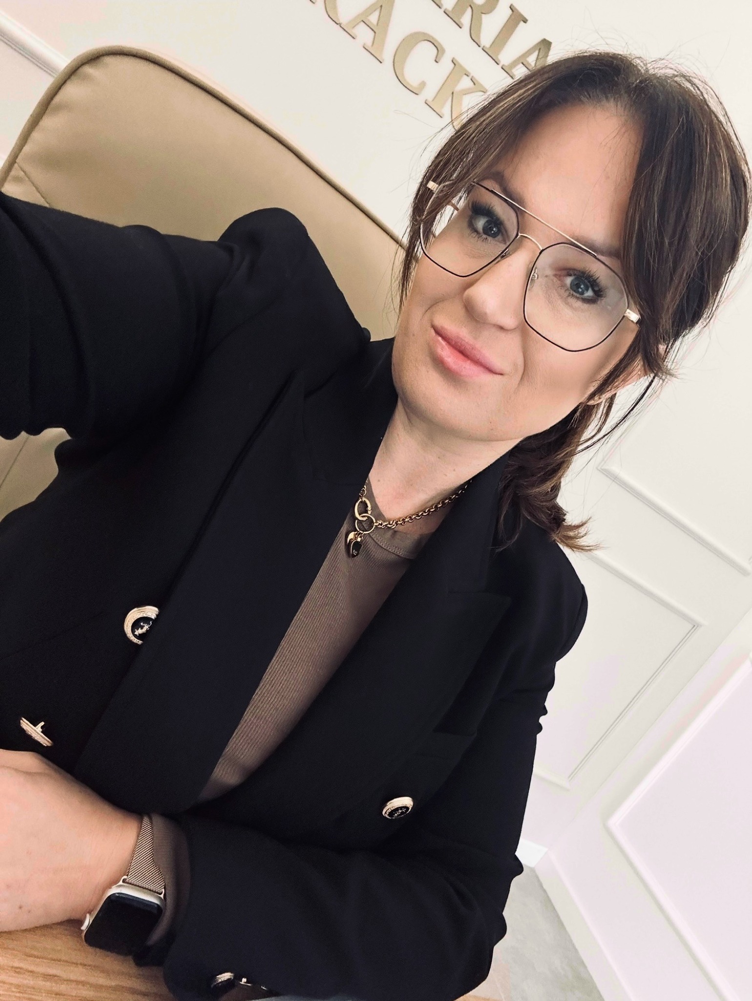 Portret kobiety w okularach i czarnej marynarce, siedzącej na krześle w biurze w Głogowie. Widoczny zegarek na ręce i złoty łańcuszek z zawieszkami.
