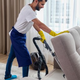 IWL - Firma sprzątająca Katowice - Mężczyzna w niebieskim fartuchu i żółtych rękawicach odkurza jasną tapicerkę sofy odkurzaczem z wężem, w tle widoczny odkurzacz i firanka w oknie.