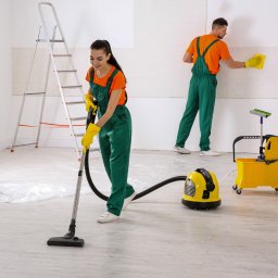 IWL - Firma sprzątająca Katowice - Dwie osoby w zielonych kombinezonach i żółtych rękawicach: kobieta odkurza podłogę, mężczyzna czyści ścianę żółtą ścierką. Podłoga zabezpieczona folią ochronną, w tle drabina i żółty wózek ze...