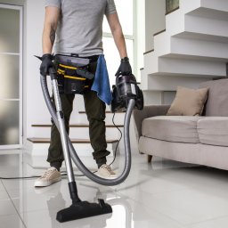 IWL - Firma sprzątająca Katowice - Mężczyzna w szarej koszulce i roboczych spodniach, z pasem narzędziowym i niebieską ściereczką, trzyma odkurzacz i rurę odkurzacza na tle białych płytek podłogowych, sofy i schodów w jasnym wnętrzu.